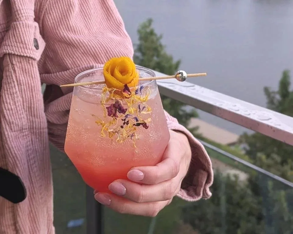 A person in a pink corduroy jacket holding a large glass of pink cocktail garnished with a rose-shaped lemon peel and edible flower petals, with a lakeside view in the background.