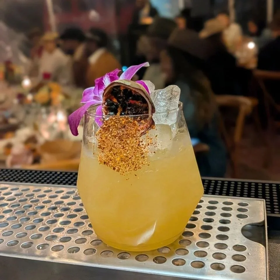 Social Butterfly ATX Rattlesnake's Kiss cocktail garnished with a dried passion fruit, fresh orchid, and chili salt rim, served at an Austin outdoor evening event with string lights