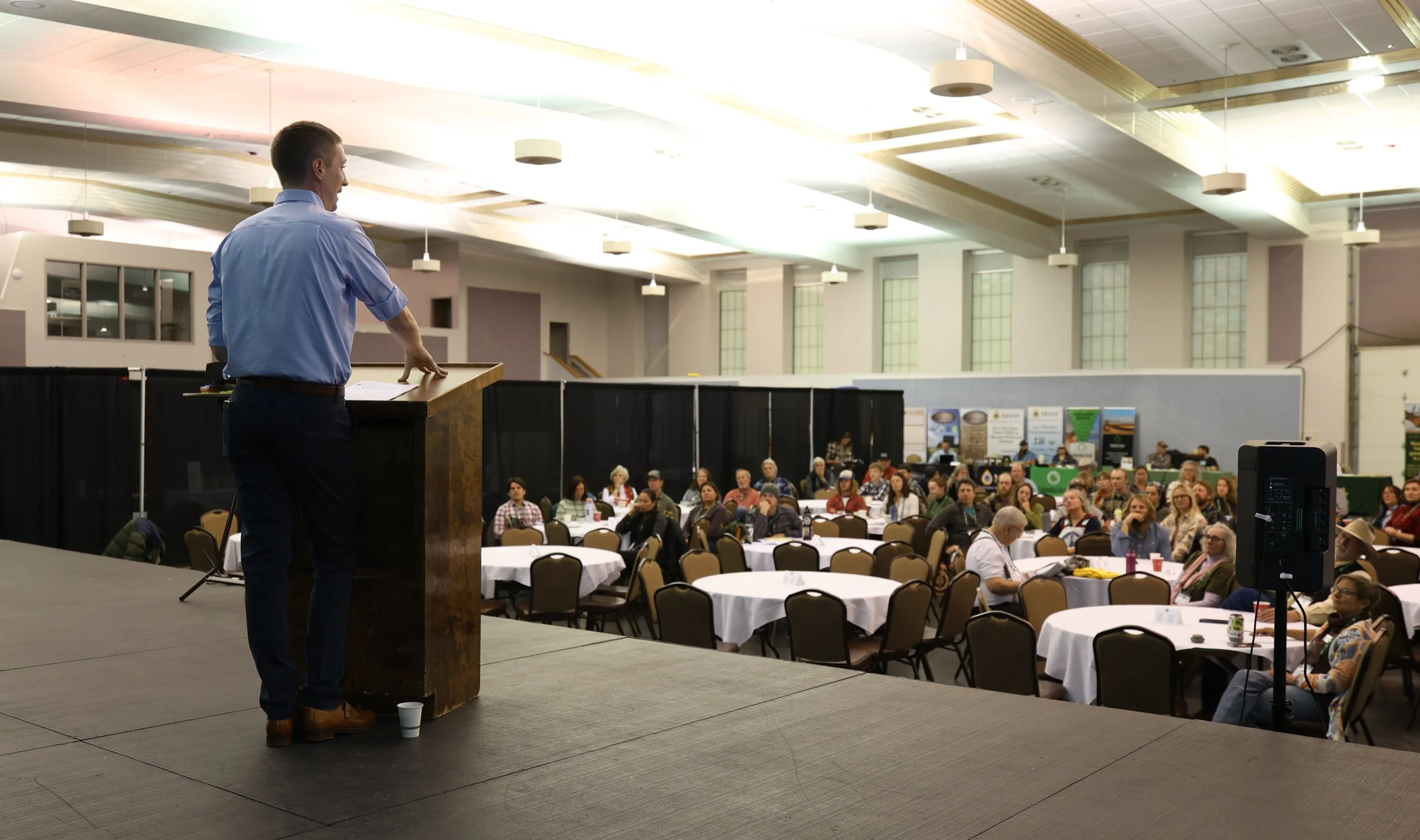 Speaker on stage at conference with audience seated at round tables in large hall