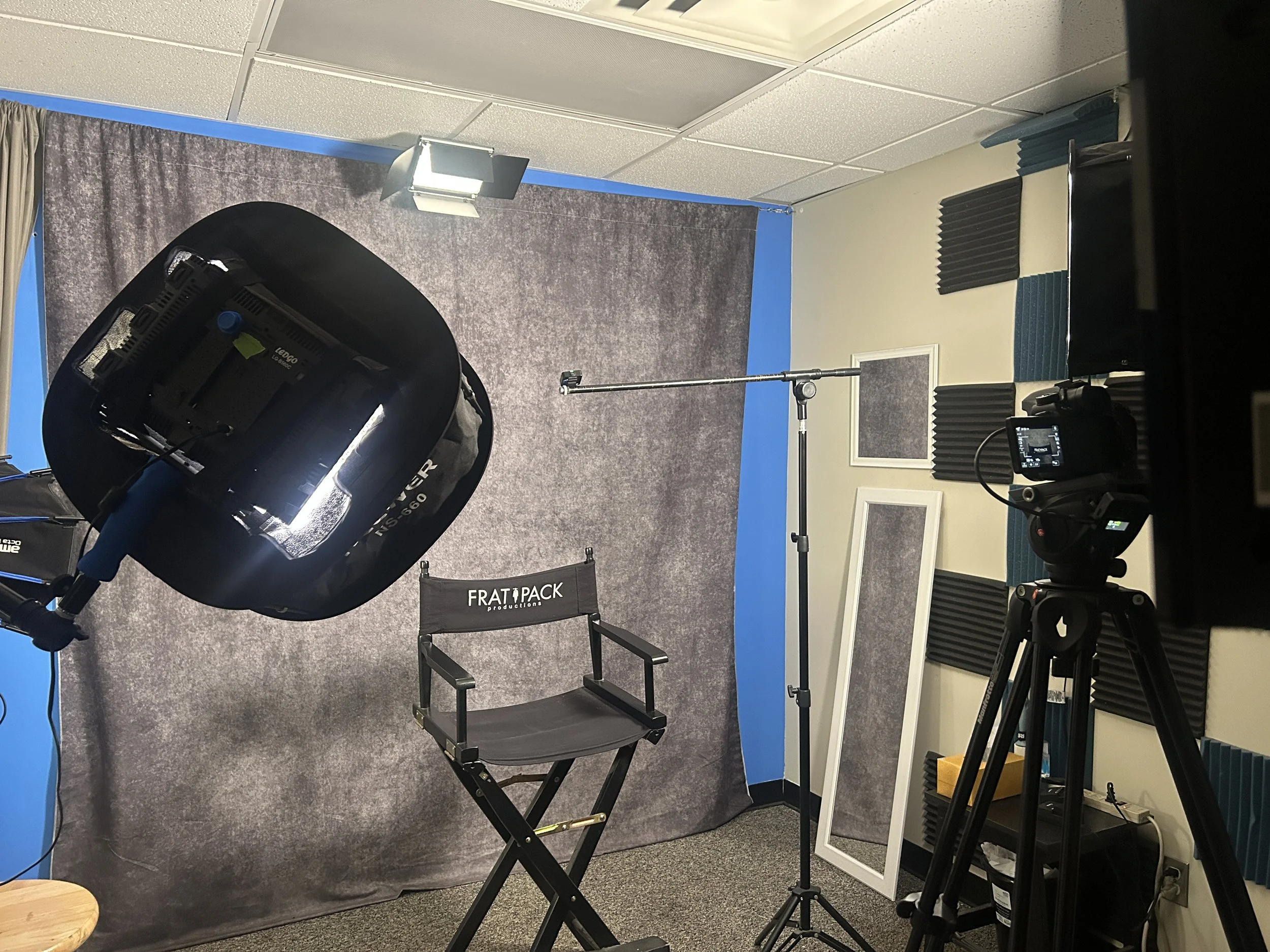 Photography studio setup with professional lighting, a camera on a tripod, a director's chair, soundproof foam panels, and a gray backdrop, indicating preparations for a photoshoot or video recording.
