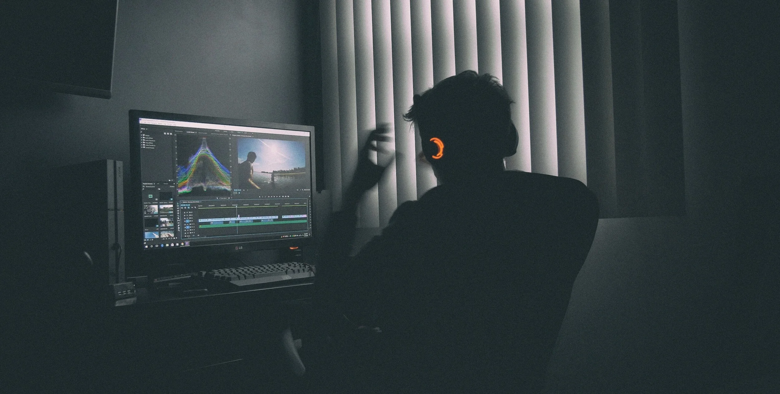 A person wearing headphones working on video editing on a computer in a dimly lit room with vertical blinds.
