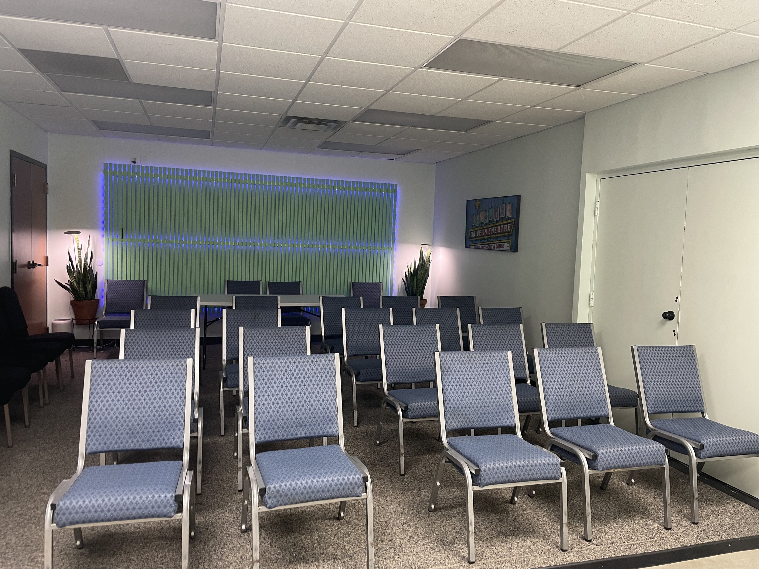 Empty chairs arranged in rows in a conference room with a decorative wall and framed poster, plants, and a closed door.