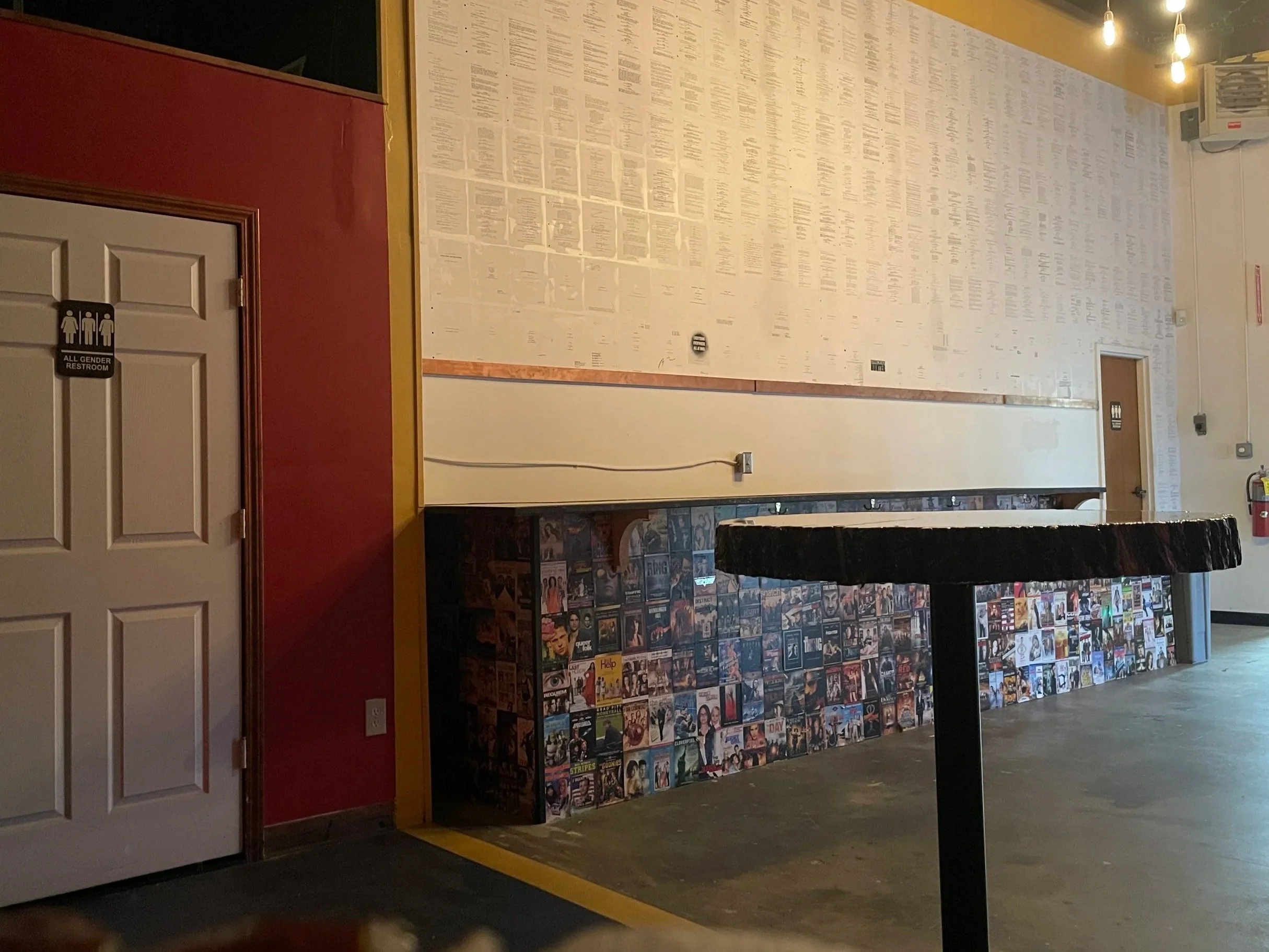 Wall covered with numerous posters and magazine covers, with a door labeled as 'All Gender Restroom' in the foreground.