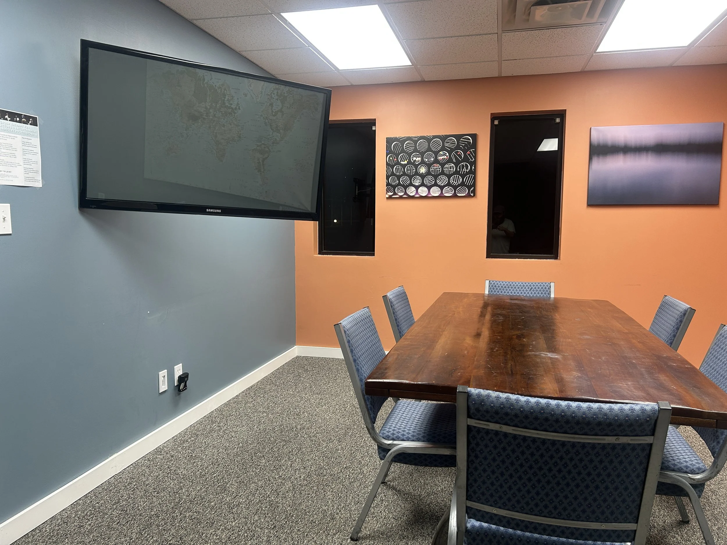 A conference room with a wooden table, six upholstered chairs, a large wall-mounted television, and framed artwork on the walls. The room has a blue and orange accent wall with a window and ceiling fluorescent lights.