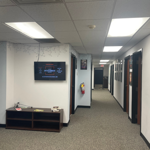 Empty office hallway with a wall-mounted TV displaying a presentation, fire extinguisher, and a black console table with papers and a pen, in an office building.