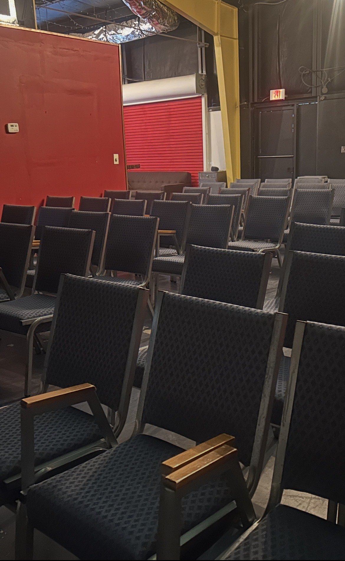 Empty auditorium with rows of black upholstered chairs with wooden armrests, a red and yellow wall with a red rolling door in the background, and a black door with an illuminated exit sign.
