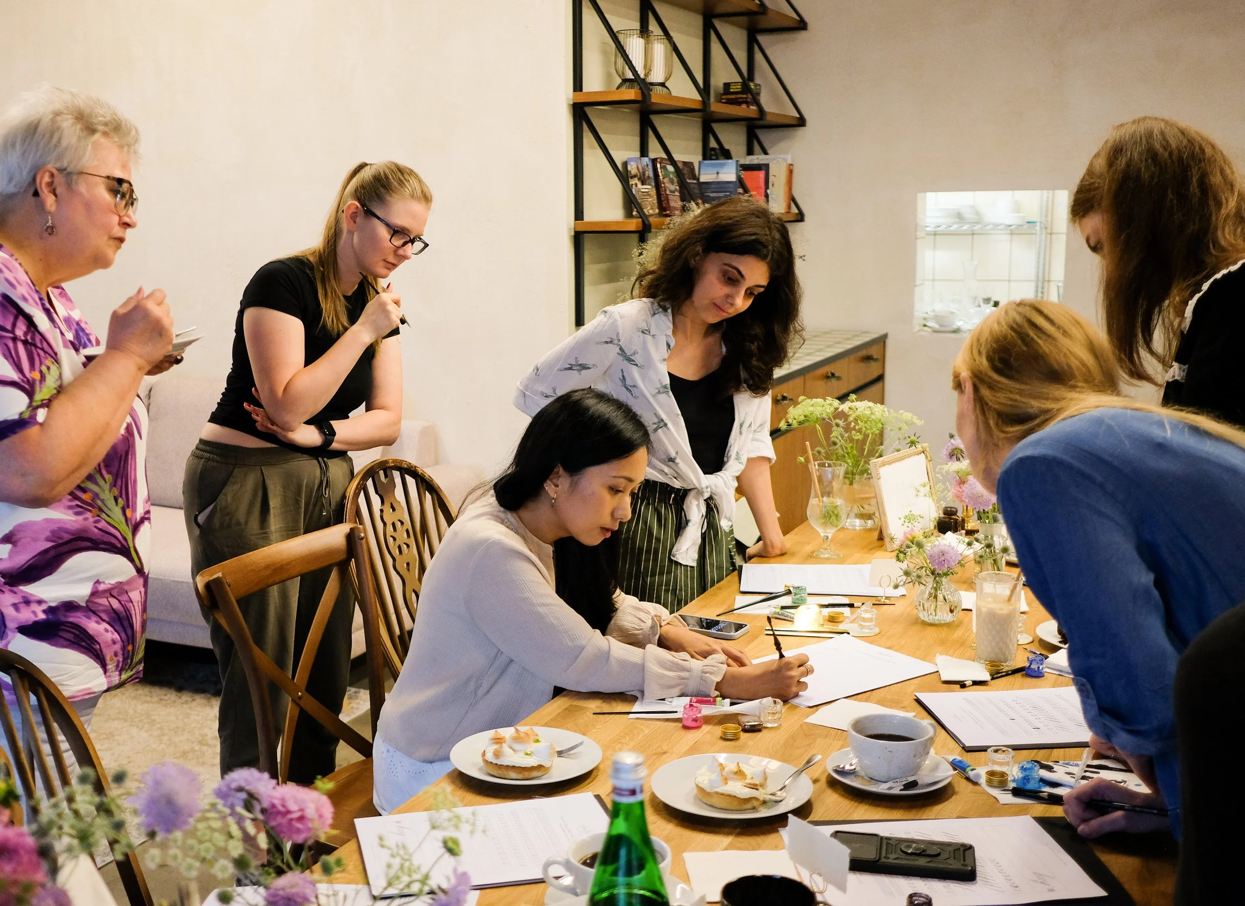 Group of women gathered around a table with notes, drinks, and desserts, engaged in a discussion or meeting in a cozy room decorated with flowers and books.