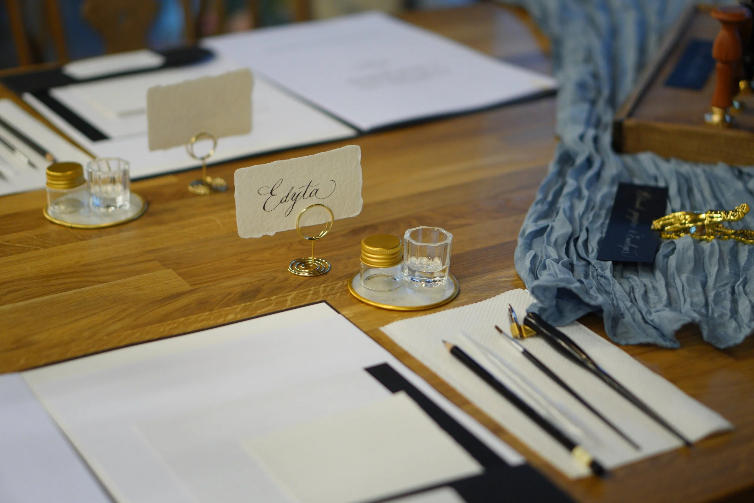 A wooden table set with placemats, pens, and glassware, featuring a small card with elegant calligraphy and a ring holder, part of a formal event or wedding decoration.