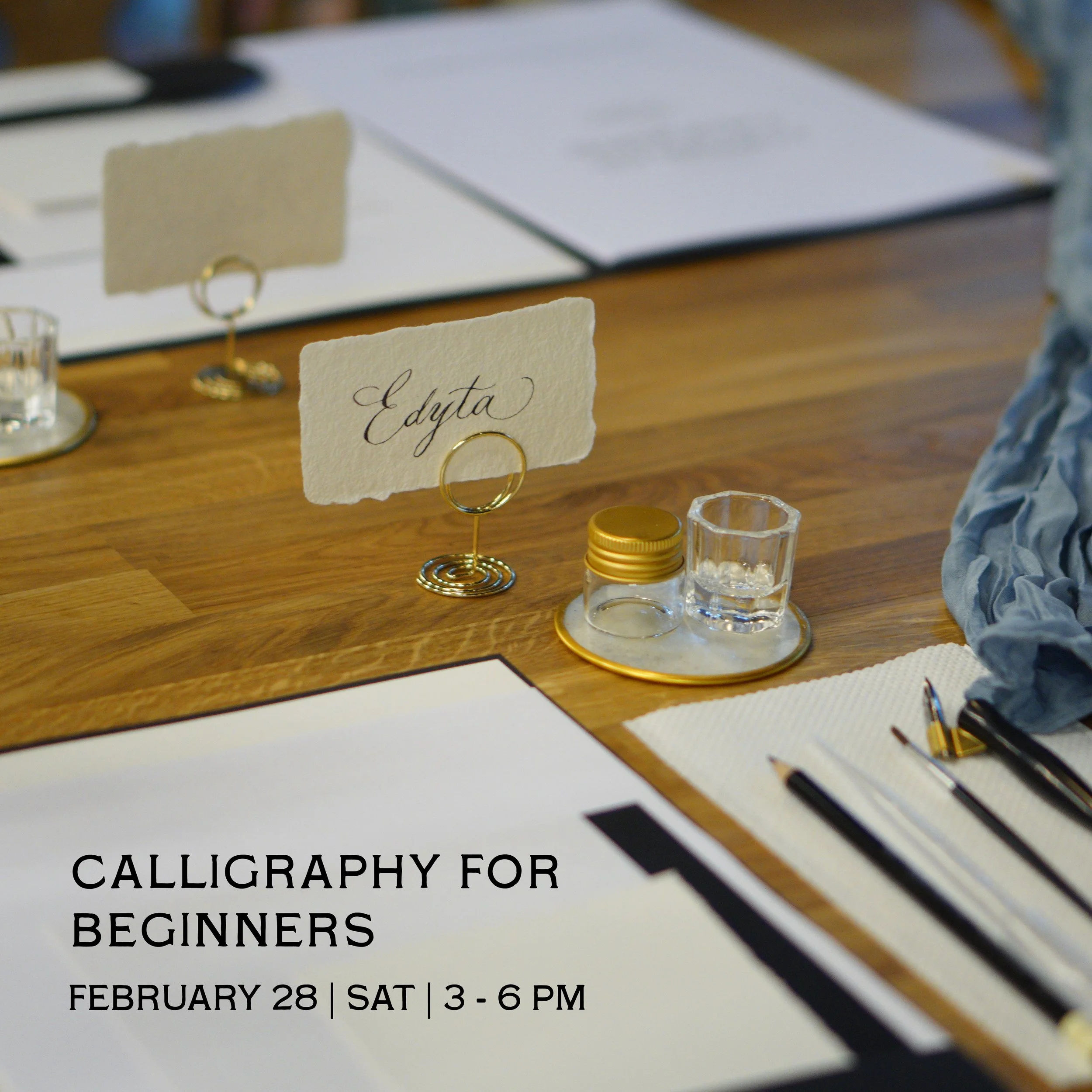 A table with calligraphy practice materials, including a sheet with writing, pens, and a place card with the name 'Edyta' on it, promoting a beginner calligraphy workshop on February 28 from 3 pm to 6 pm in Krakow Poland.