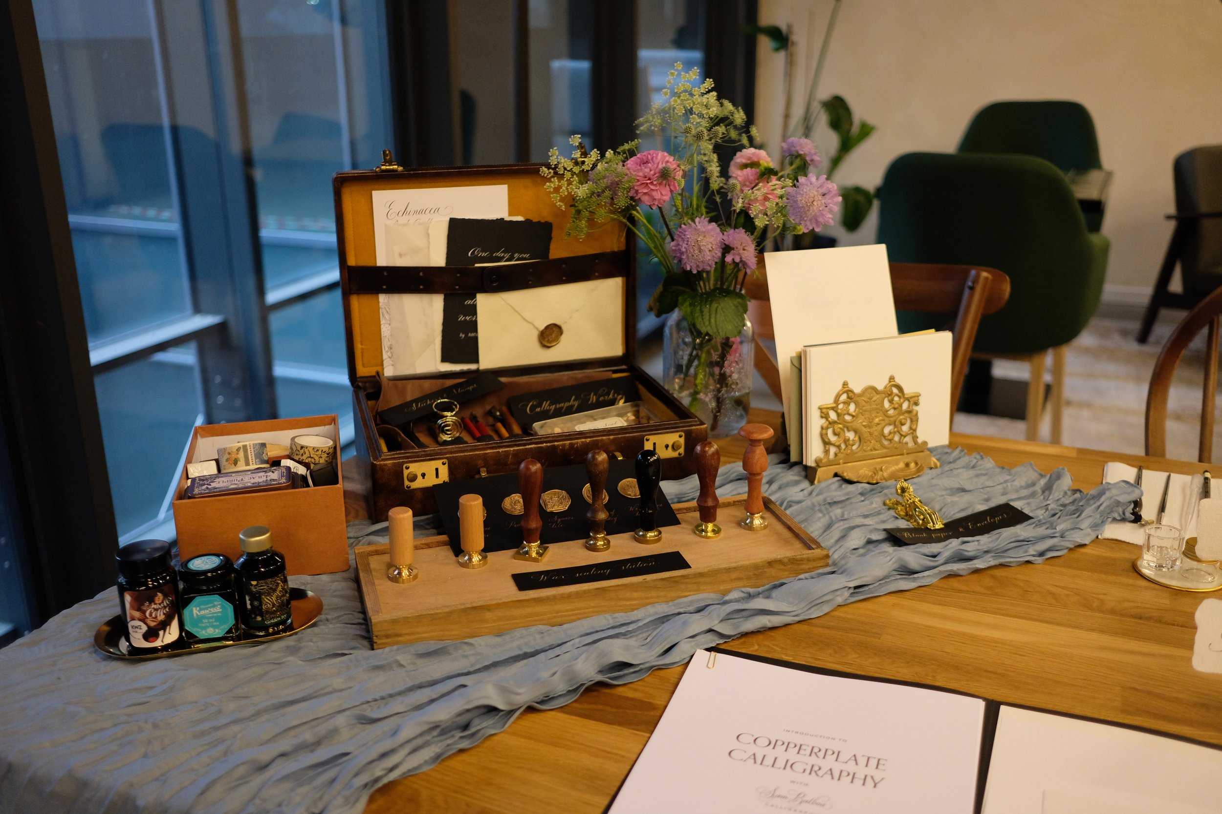 Table display with calligraphy supplies, ink bottles, stamps, wax seals, a flower arrangement, and decorative items in a well-lit room.