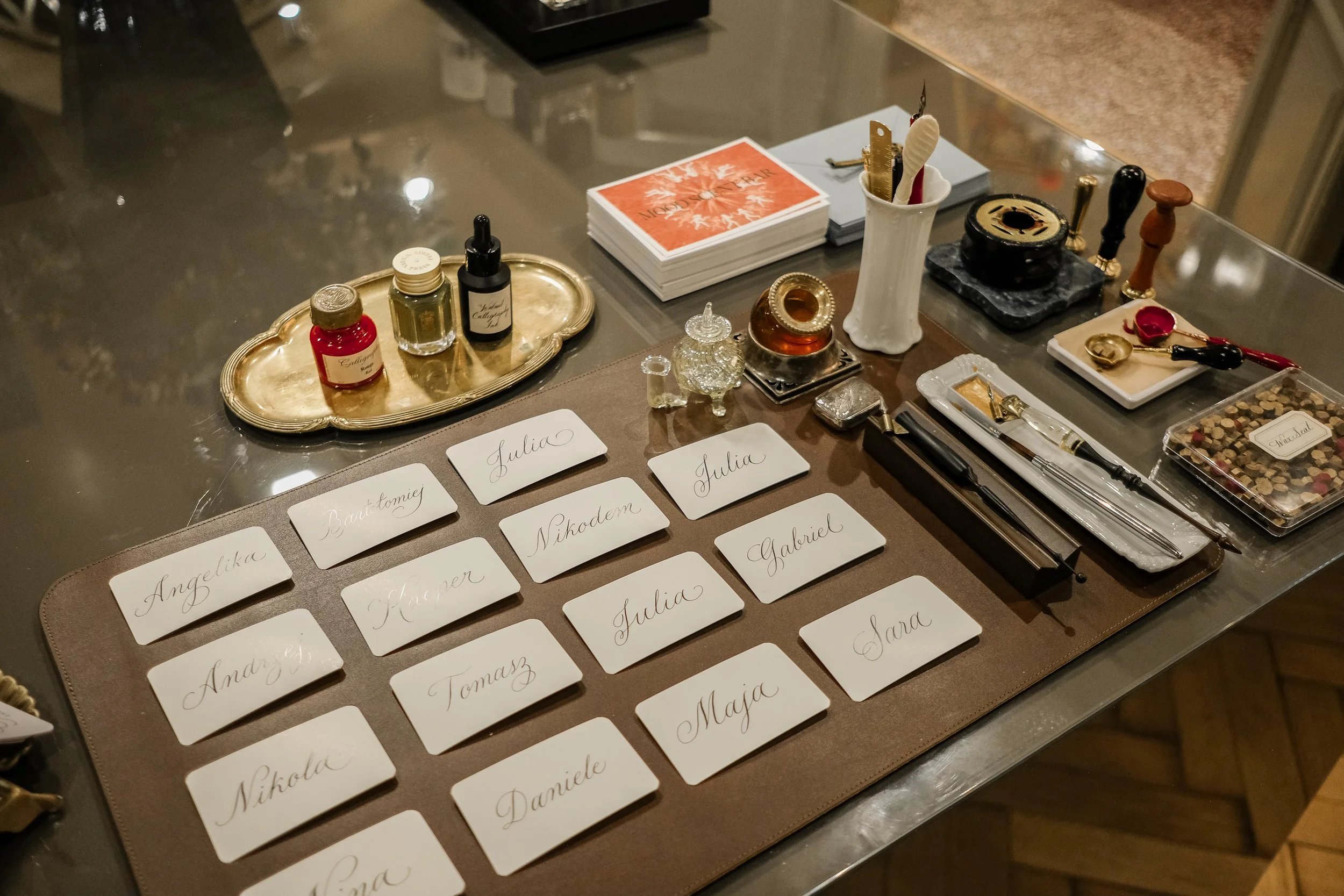 Name tags with handwritten cursive names arranged on a brown leather sheet. Items for a formal event, including ink bottles, pens, and other table accessories, are on the table behind.