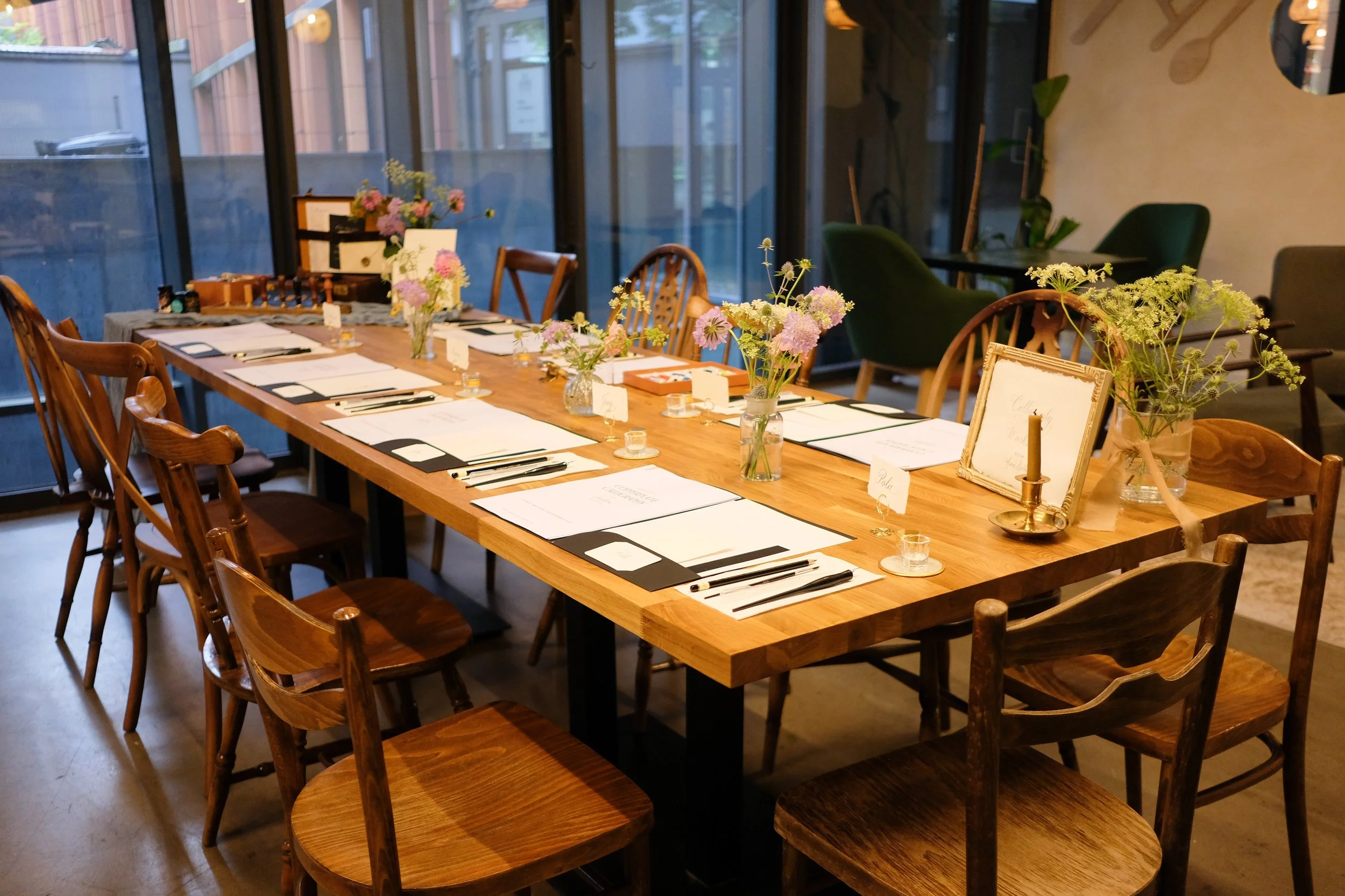 A long wooden table set for a creative calligraphy workshop with floral centerpieces and table settings, located in a cozy, stylish restaurant with large glass windows.