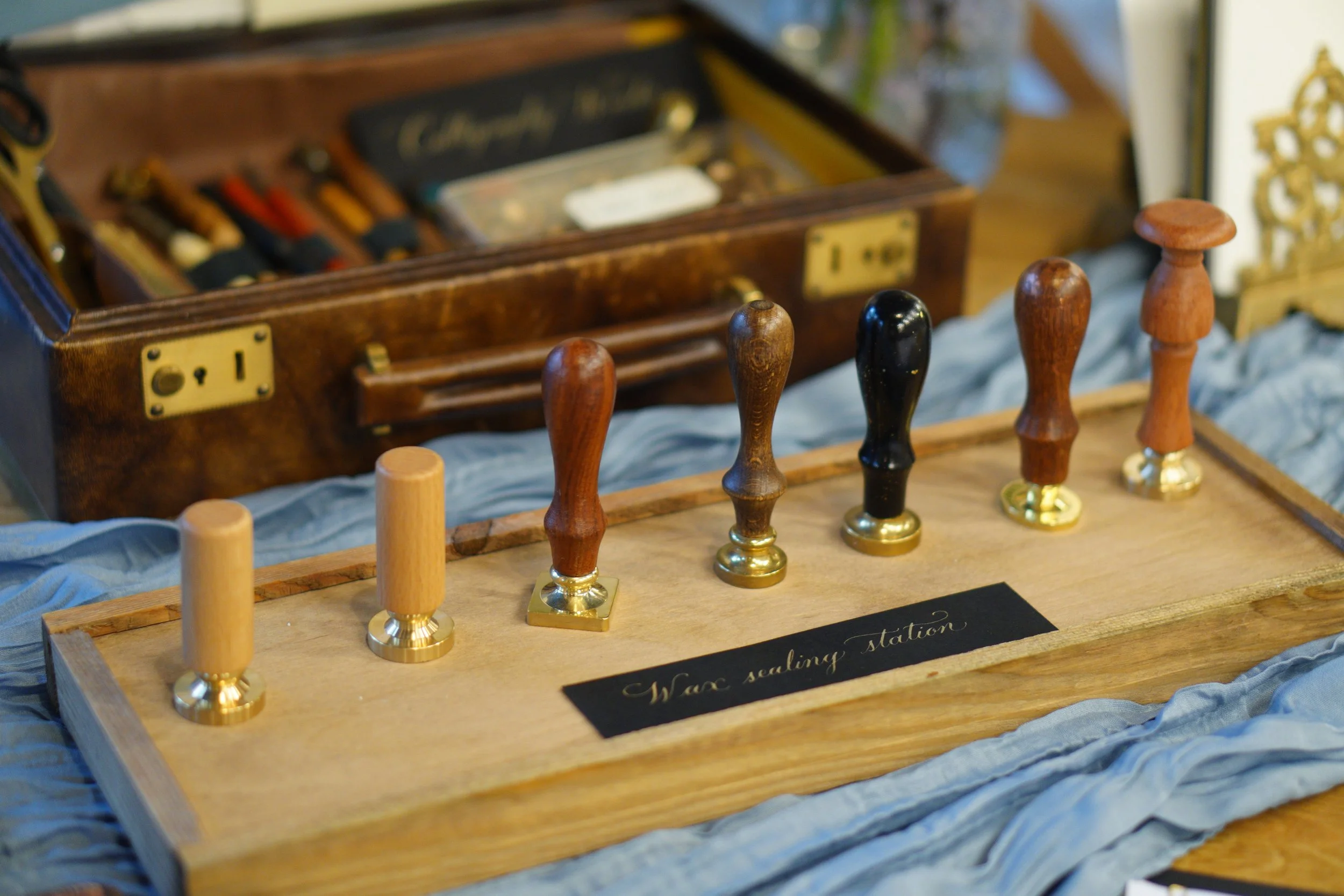 A wax sealing station with six wax seal stamps of different designs and wood handles, arranged on a wooden tray. A black plaque with gold cursive writing reads 'Wax sealing station.' In the background, there is a wooden box with various tools and sup
