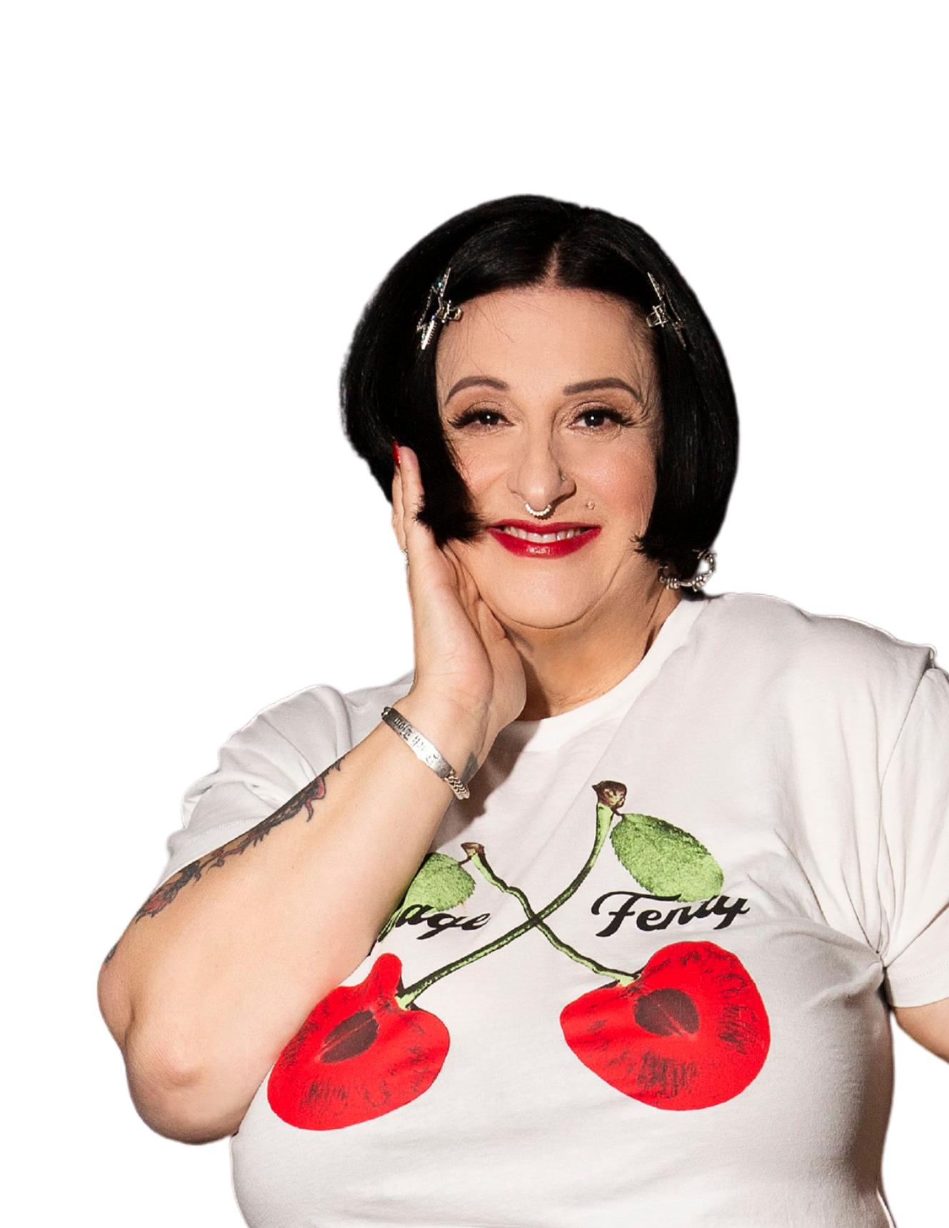 Woman with short black hair, red lipstick, and tattoos, wearing a white T-shirt with cherries and the word "Fury" on it, smiling and posing with her hand on her face.