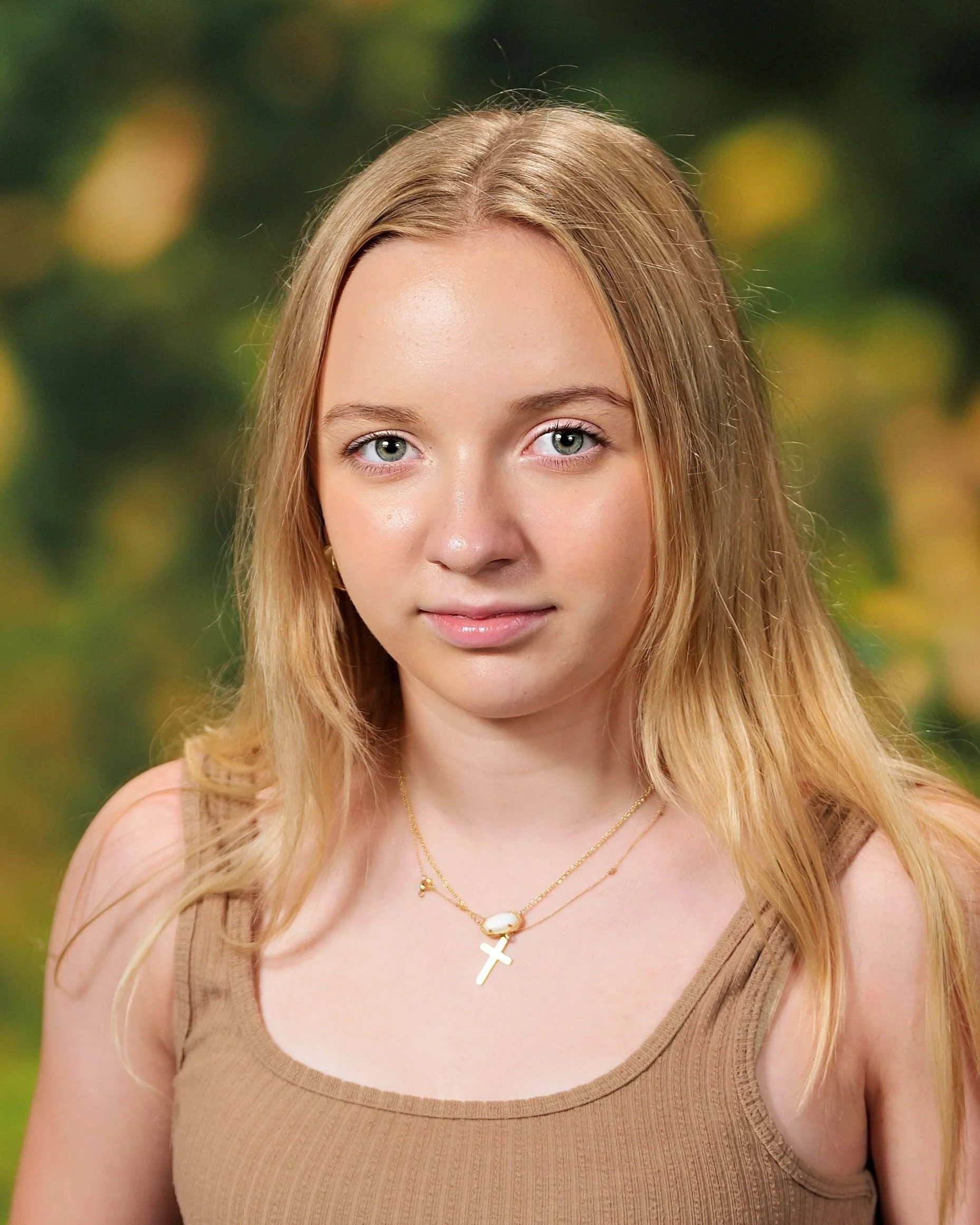 Portrait of a young girl with long blonde hair, wearing a pink sweater and a gold necklace, outdoors with a blurred green background.