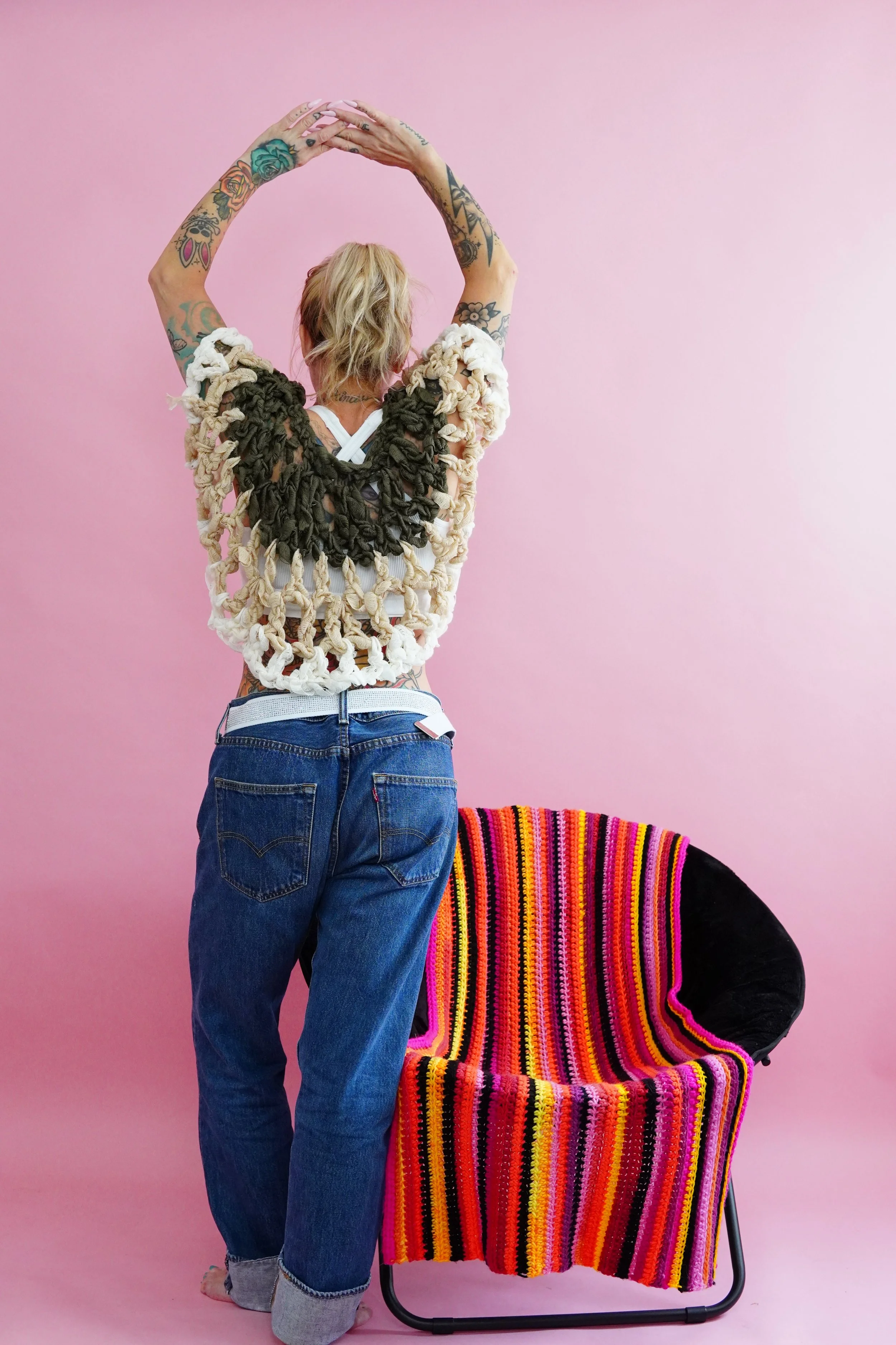 A person with tattoos on their arms, blonde hair, and wearing a knitted top, standing with their back to the camera. They are in front of a pink background, with a colorful striped chair nearby, wearing a handmade crocheted ribbon cropped cape.