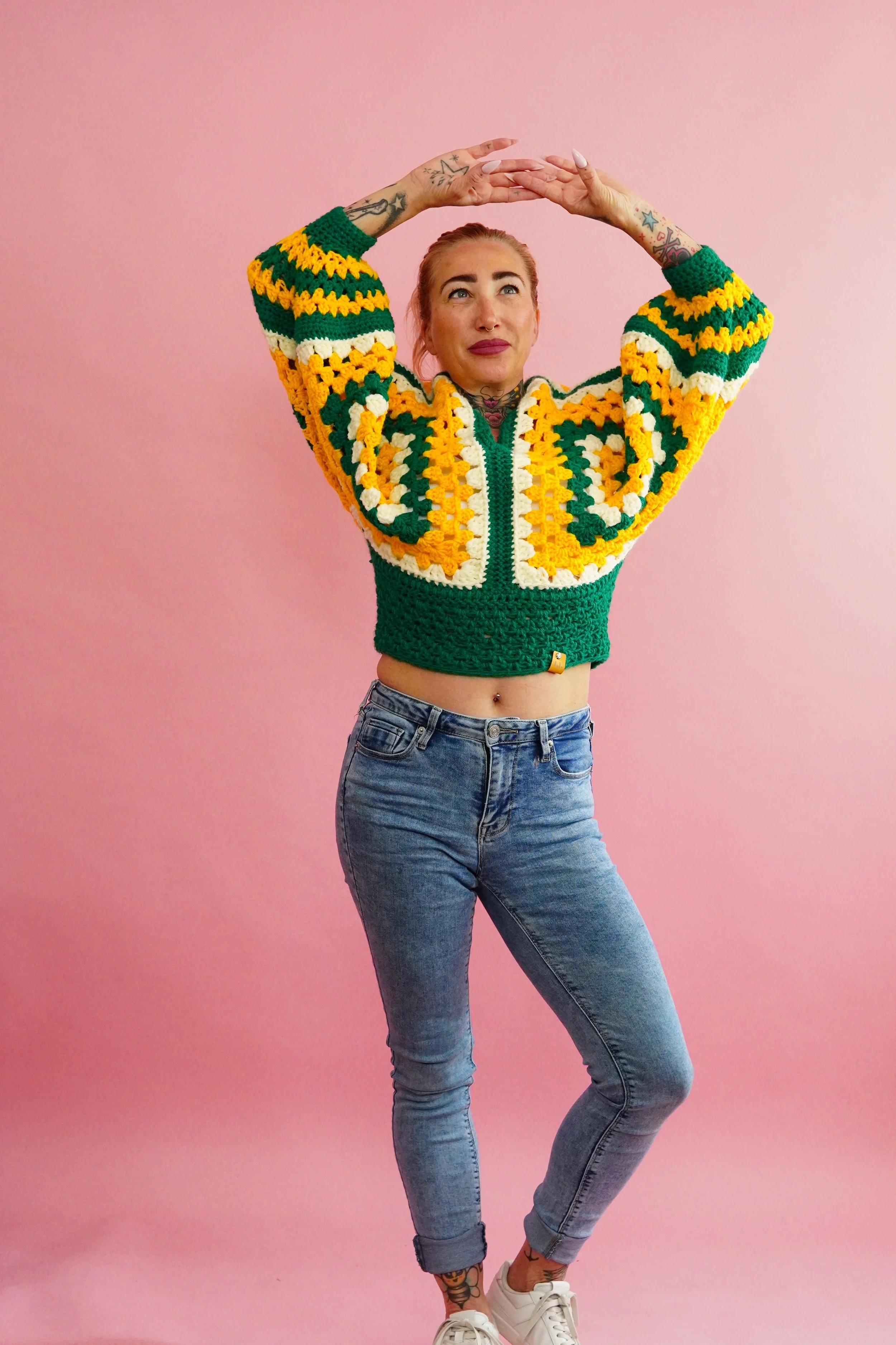 A woman standing against a pink background, wearing a colorful crochet sweater, blue jeans, and white sneakers, with tattoos visible on her arms and neck, and her hands raised above her head.