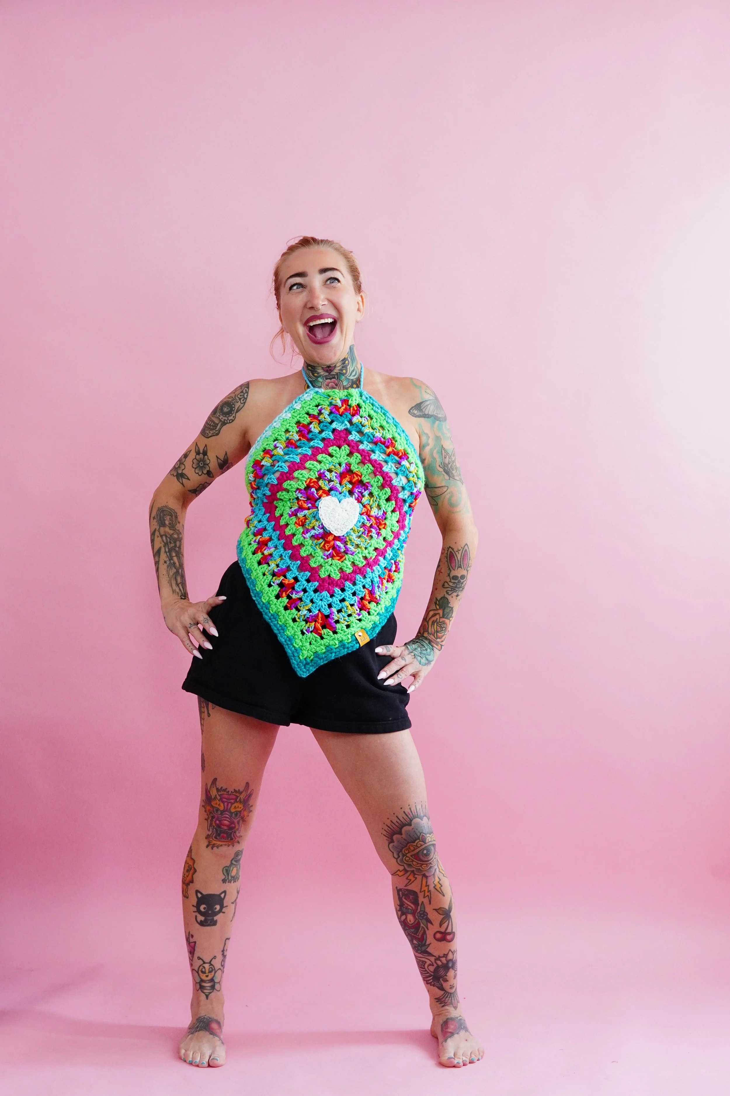 A young woman with tattoos on her arms and legs, laughing and standing against a pink background. She is wearing a colorful crocheted halter top with a heart on the front and black shorts.