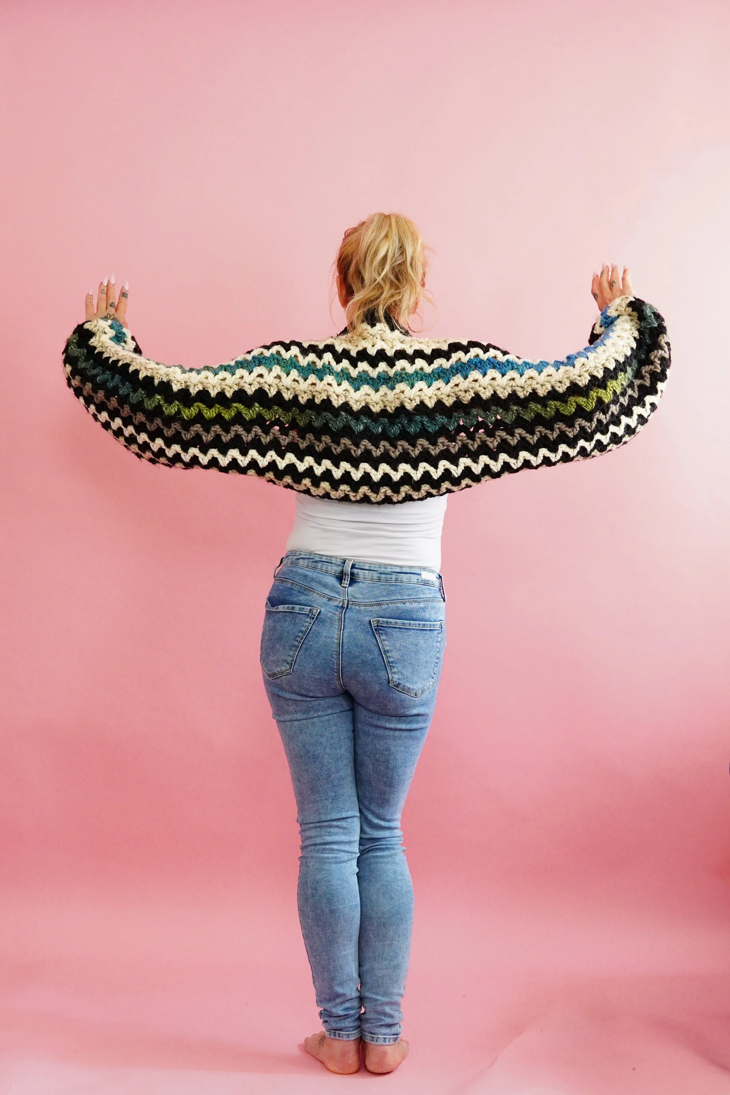 Person standing with their back to the camera, wearing a striped, multicolored crocheted shrug, white top, and jeans, against a pink background.