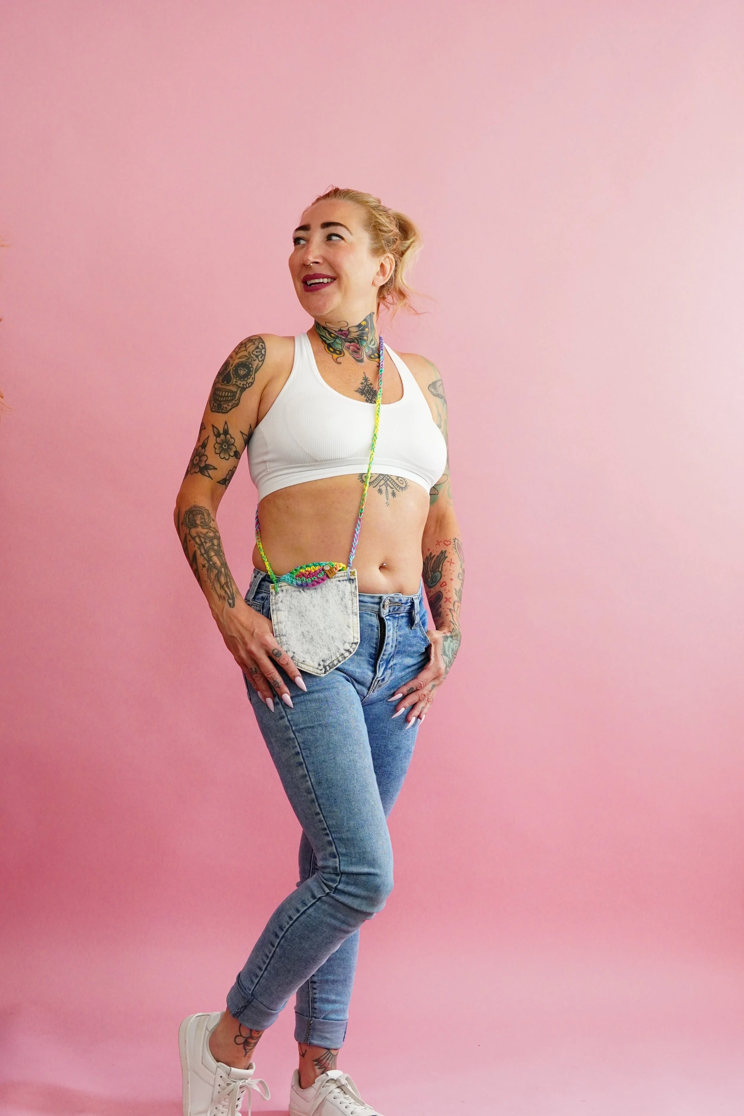 A woman with tattoos wearing handmade denim purse, a white sports bra, jeans, and white sneakers standing against a pink background.
