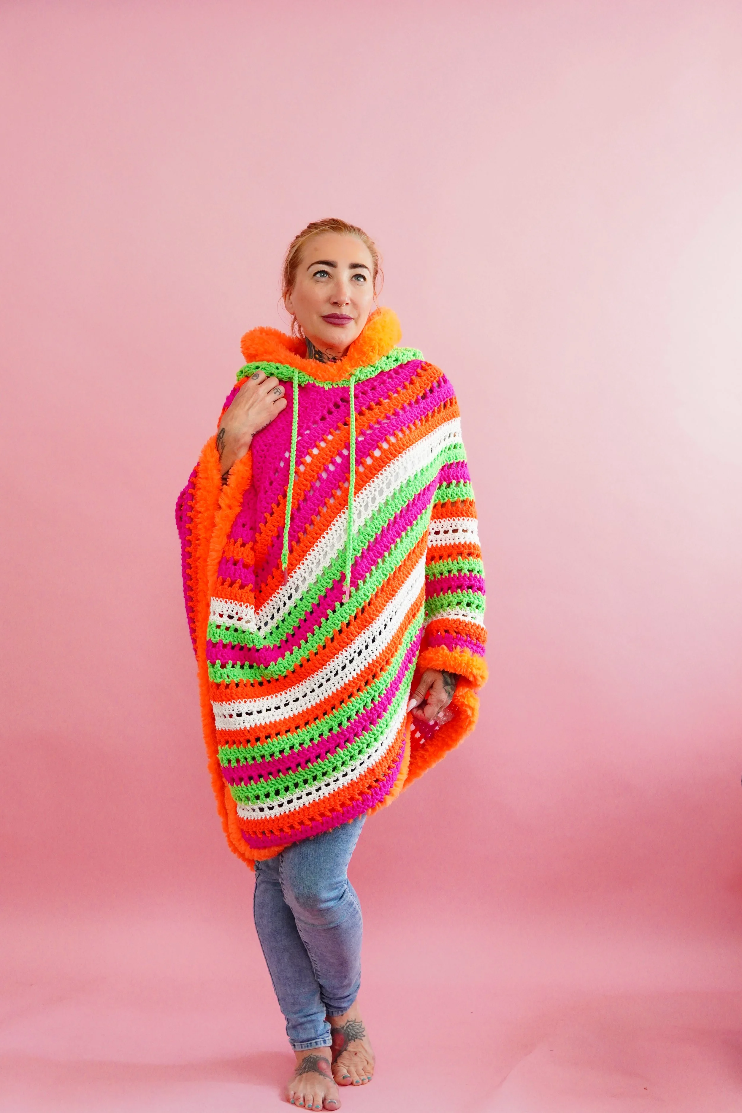 A woman with blonde hair and tattoos on her feet wearing a colorful, striped black light activated crochet poncho and standing barefoot against a pink background.