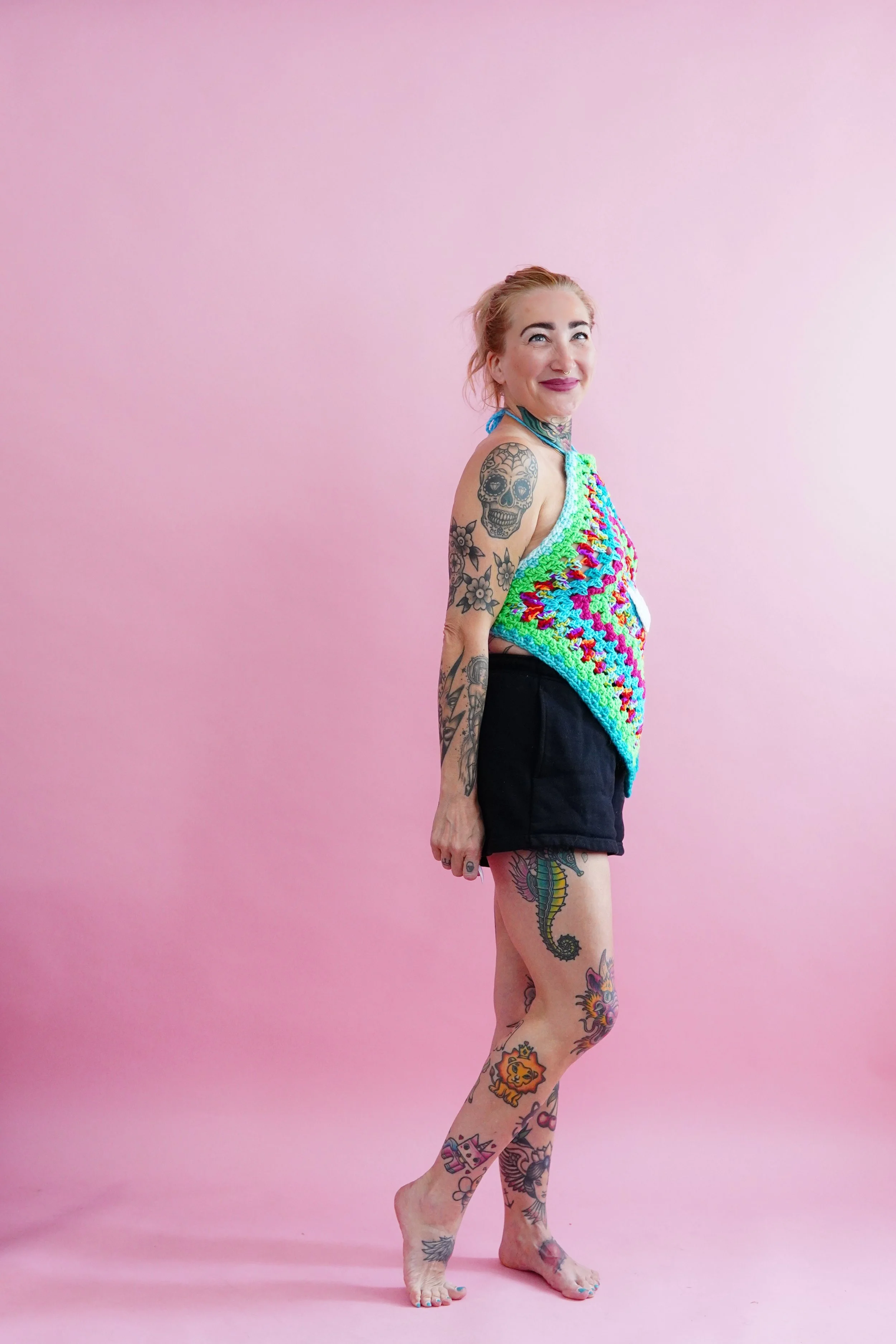 Woman with colorful tattoos standing against a pink background, wearing a crocheted summer top and black shorts, smiling at the camera.