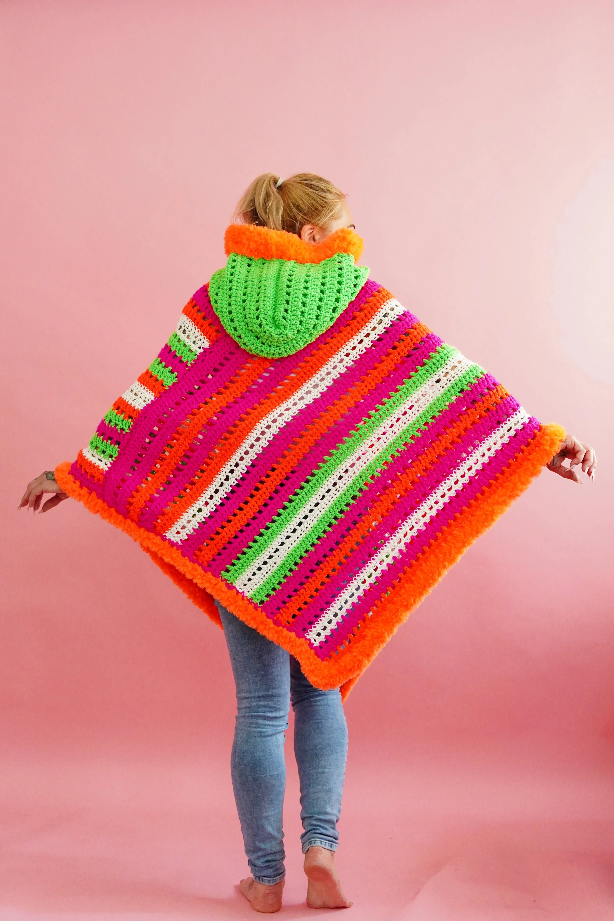 Person wearing a colorful, oversized black light activated crochet poncho with a hood, standing against a pink background.