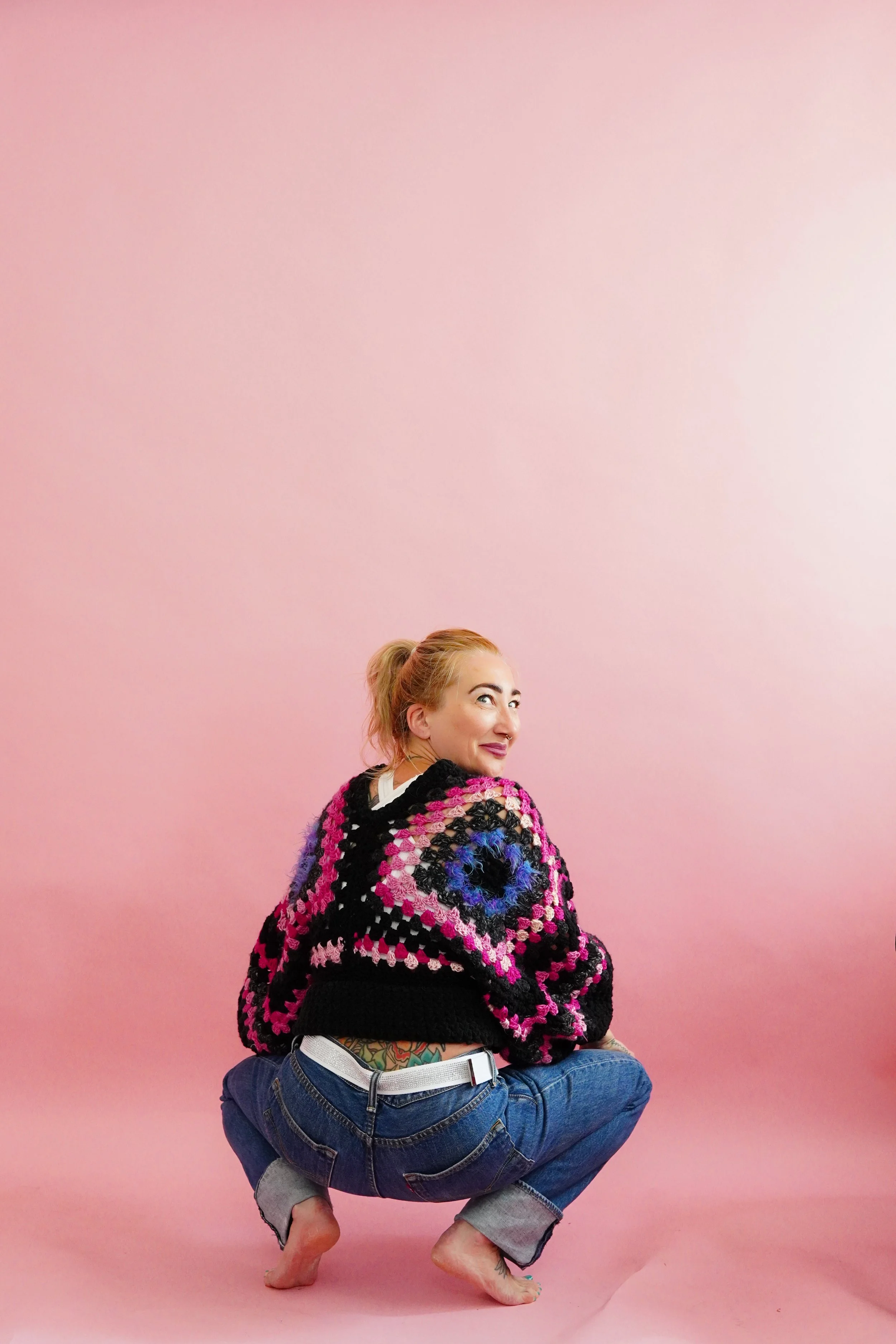 A woman with a ponytail wearing a colorful crochet sweater, blue jeans, and no shoes, squatting barefoot against a pink background, looking over her shoulder with a playful expression.