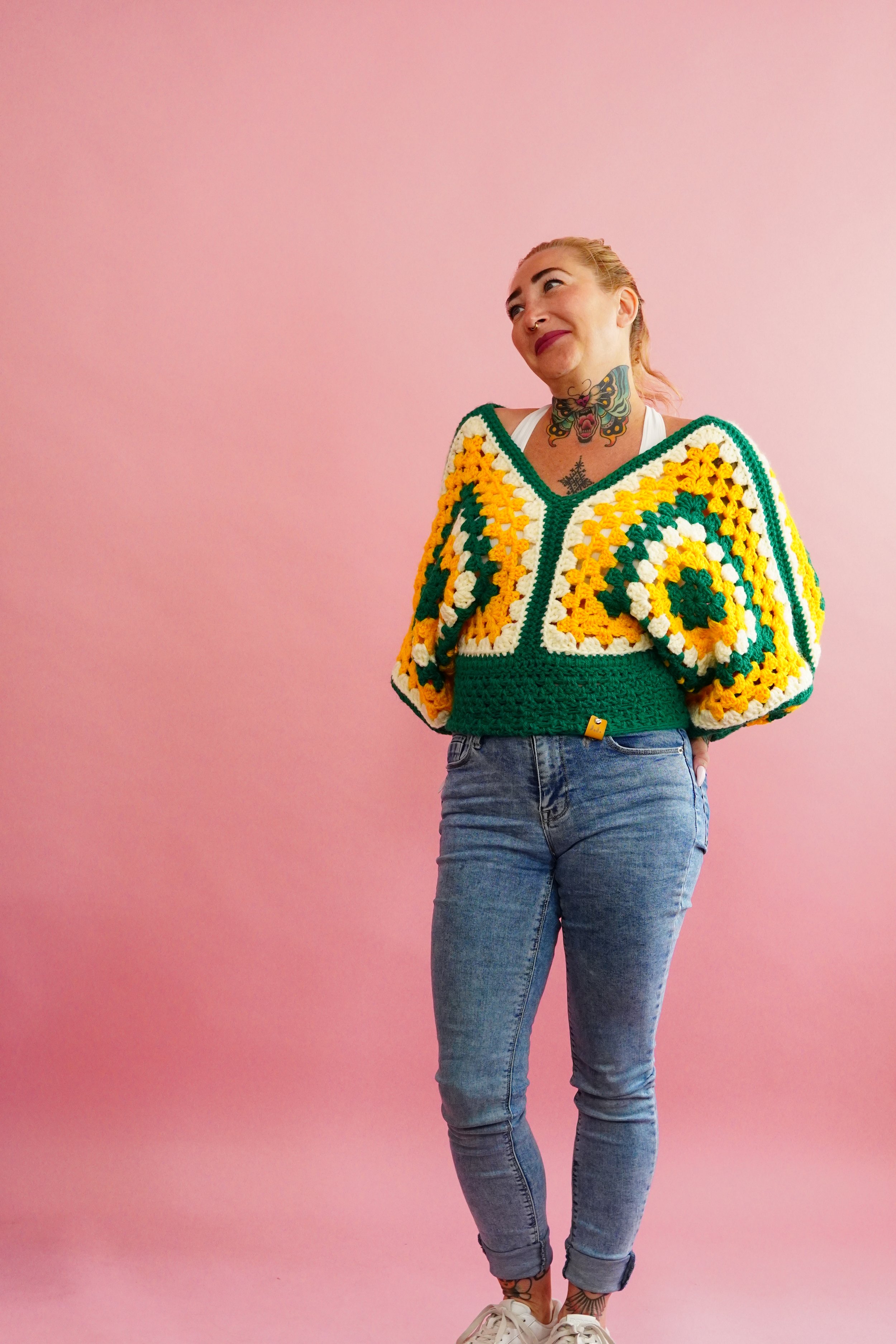 Woman with tattoos wearing a colorful crochet sweater, blue jeans, standing against pink background.