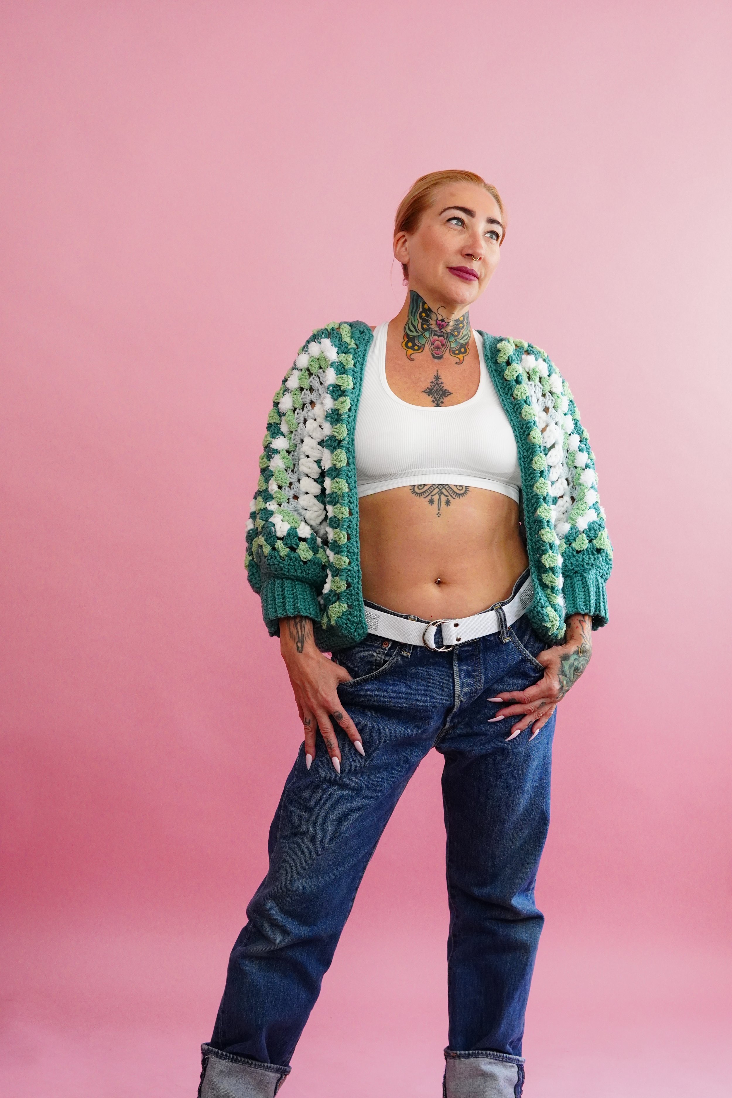 A woman with tattoos wearing a colorful crochet sweater, white sports bra, and jeans against a pink background.