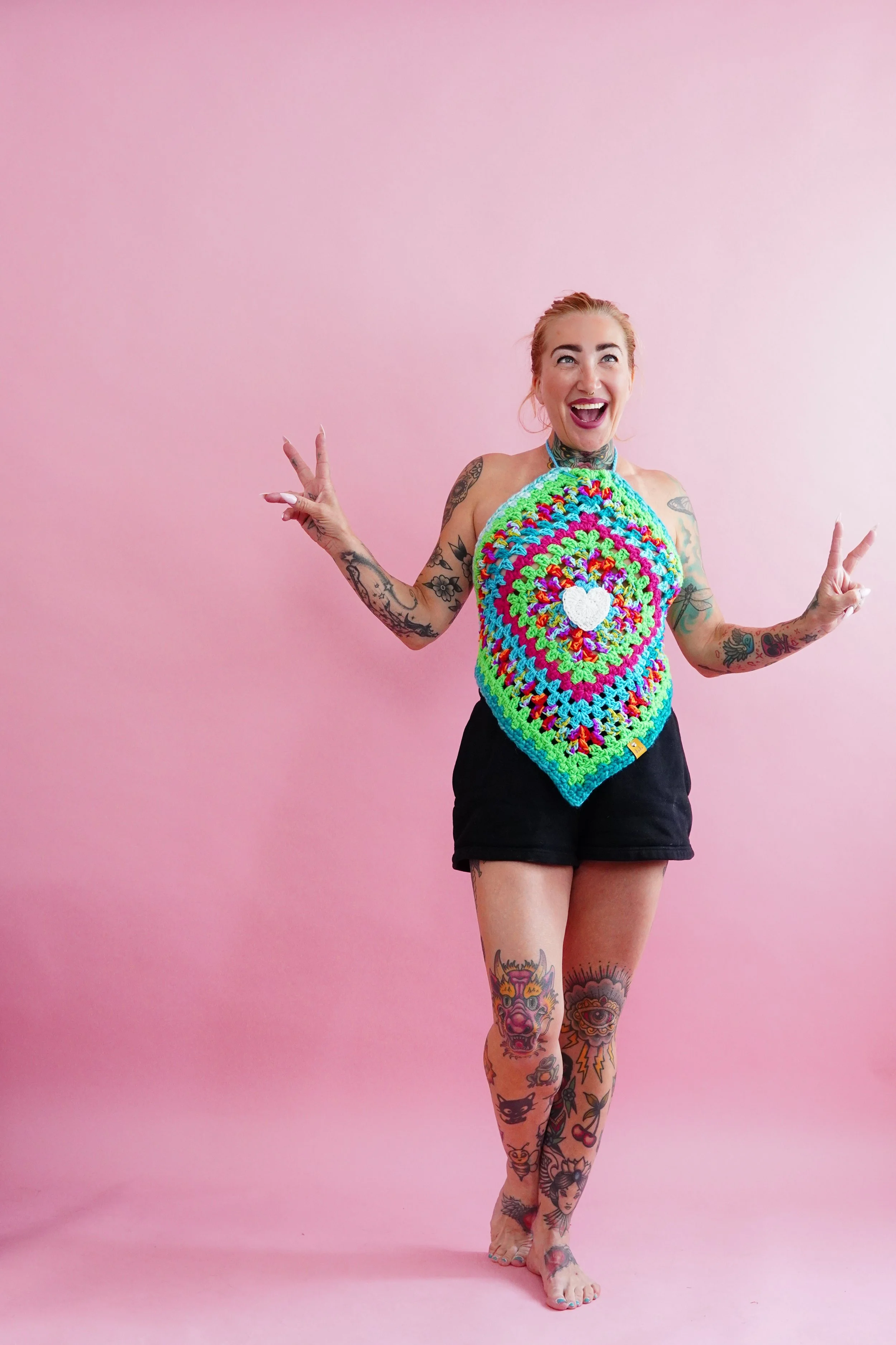 A woman with tattoos on her arms and legs, wearing a colorful crochet summer tank top and black shorts, standing against a pink background, smiling and making peace signs with her hands.