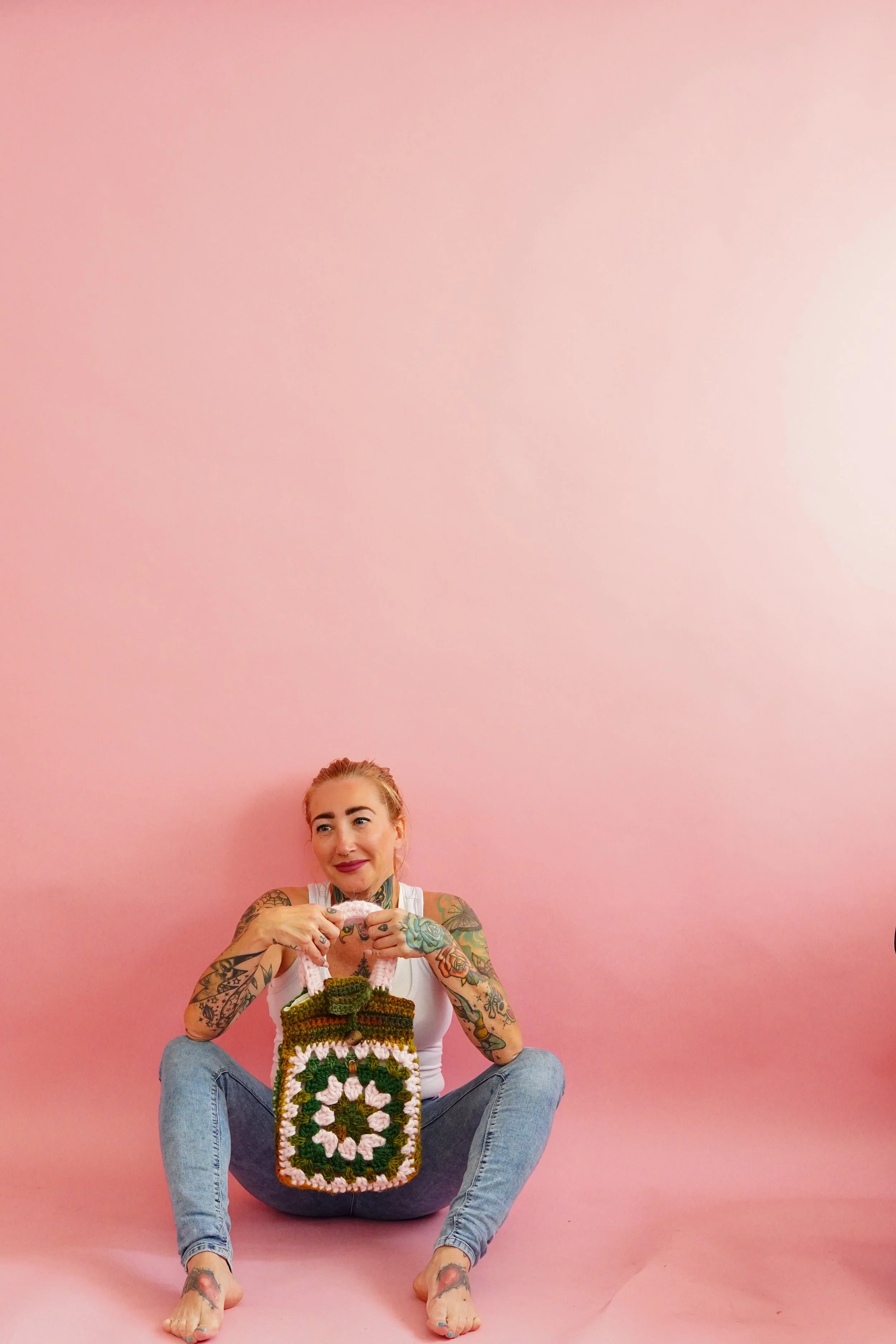 Tattooed woman sitting against a pink background, holding a colorful crocheted bag, smiling with her hair pulled back.