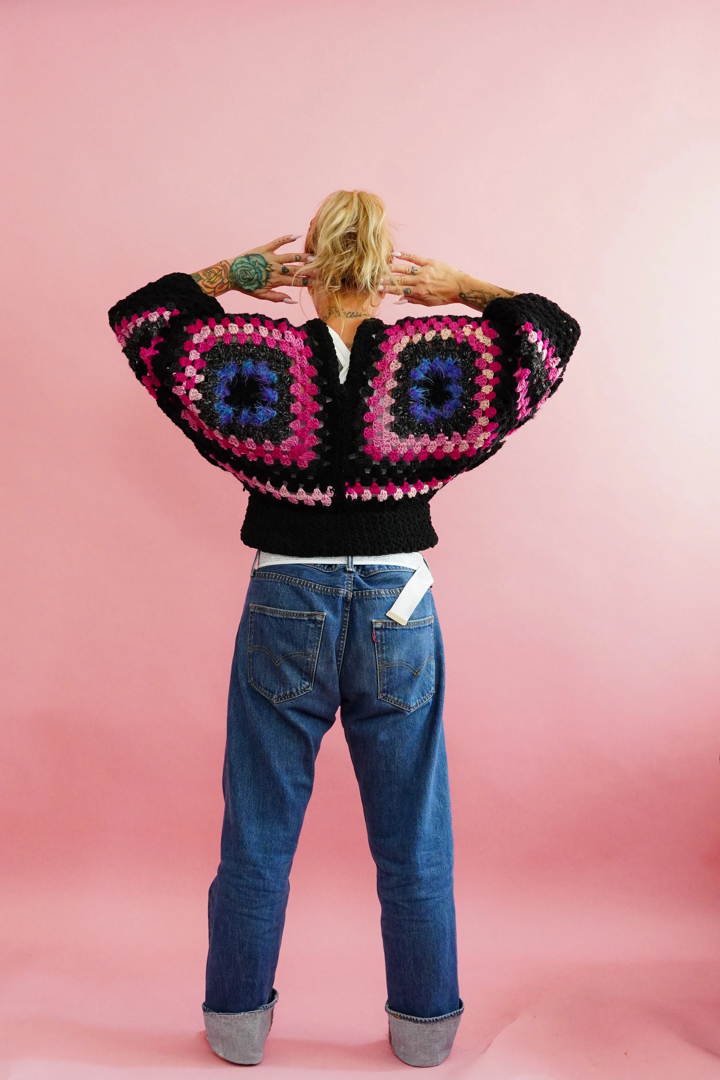 Person standing with back to camera, wearing a colorful crochet sweater with eye patterns, blue jeans, and a pink background