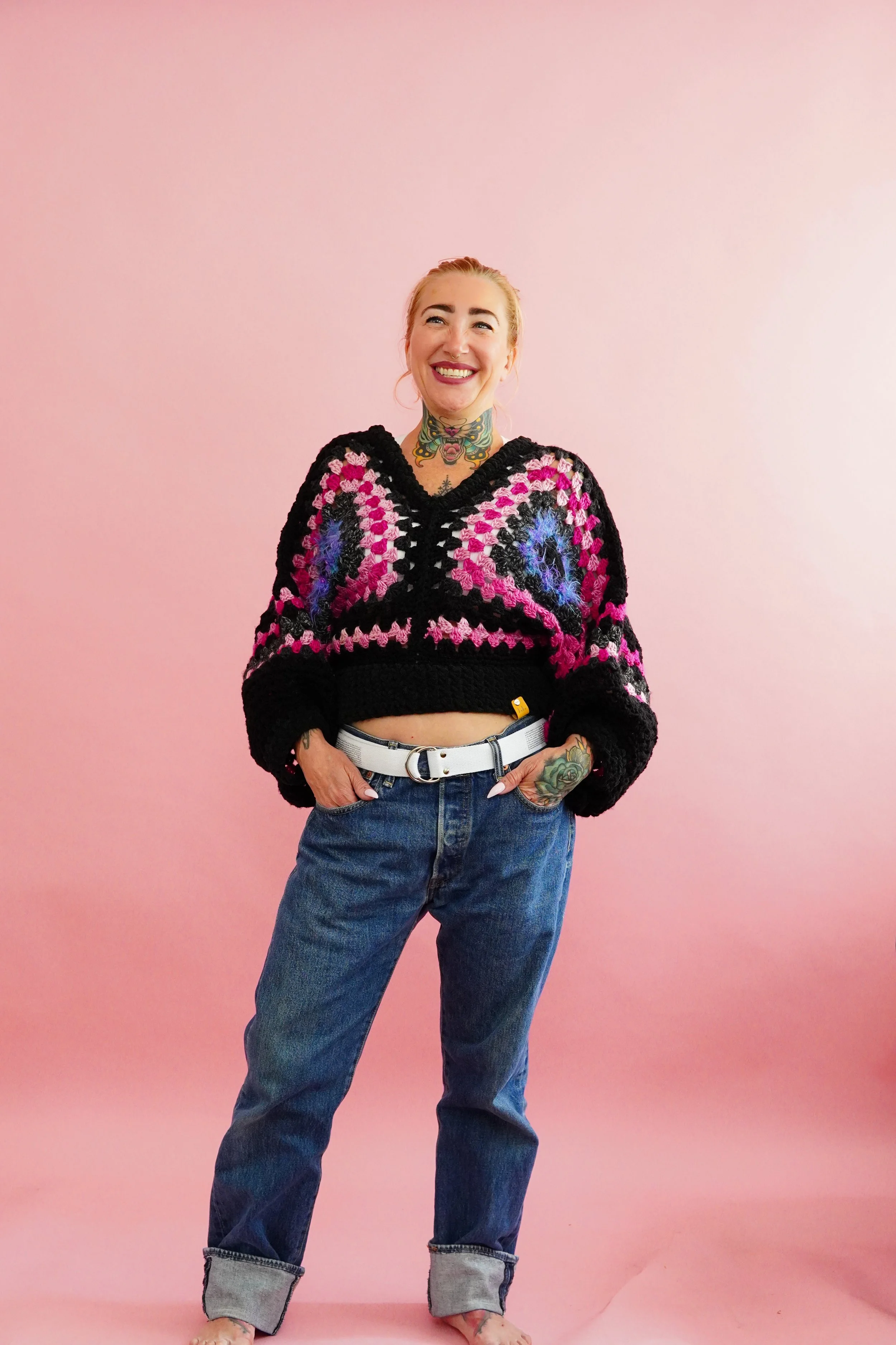 A woman with blonde hair tied back, smiling, wearing a soft black pink purple crochet sweater, blue jeans, and a white belt, standing barefoot against a pink background.
