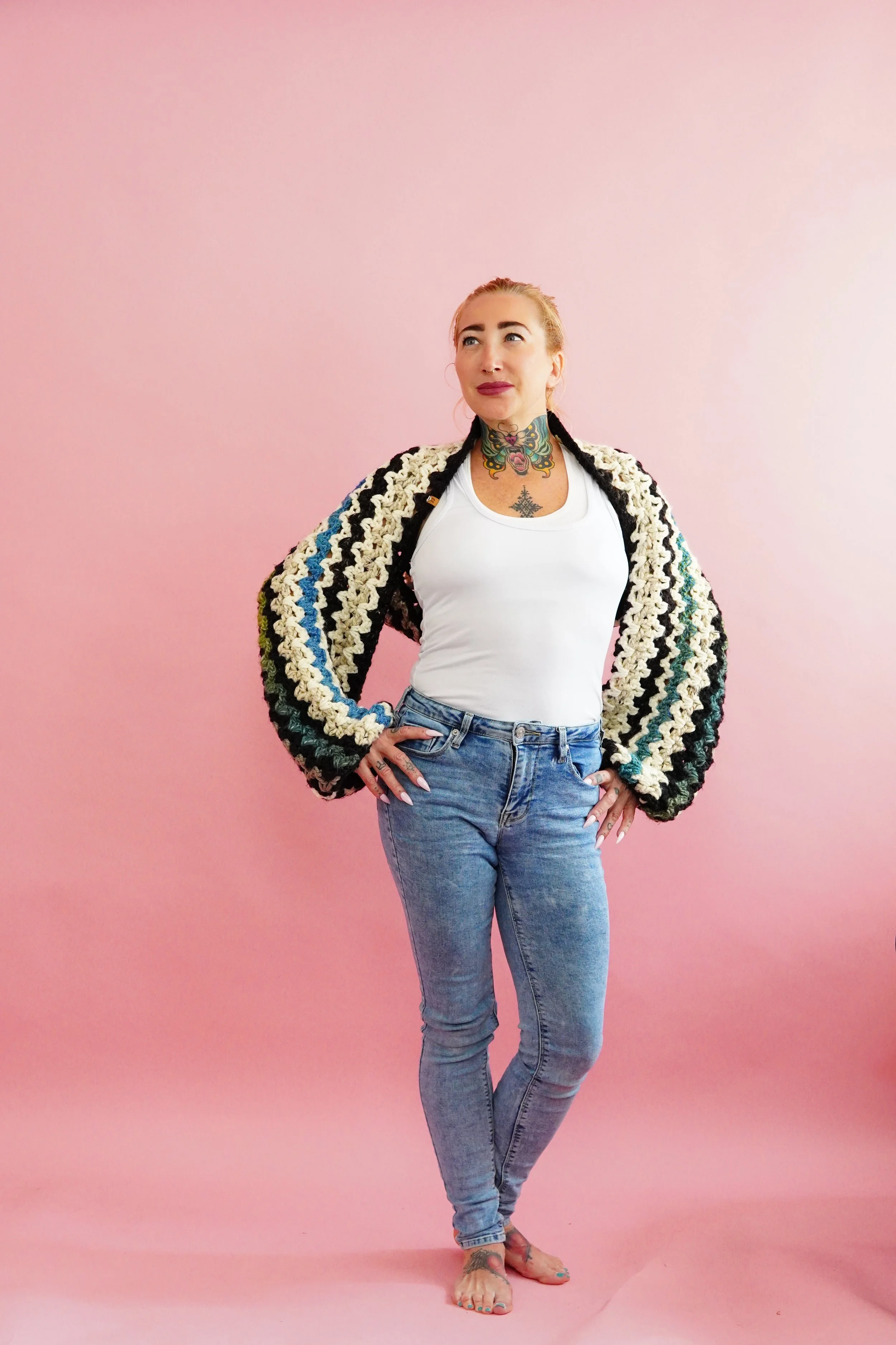 A woman standing barefoot against a pink background, with her hands on her hips, wearing a white tank top, a multicolored crocheted jacket, and blue jeans, with tattoos visible on her neck, chest, and feet.