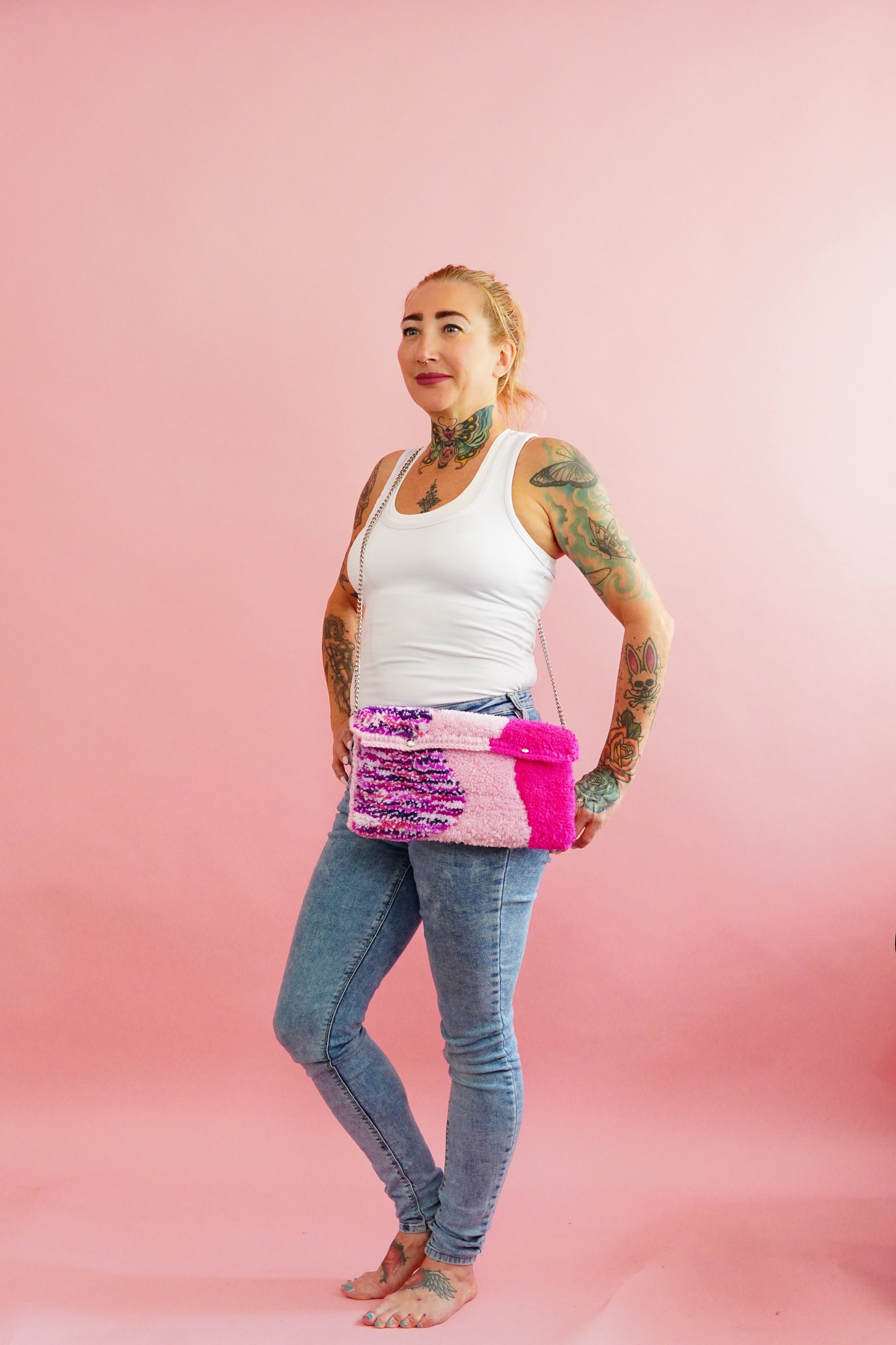 A woman with tattoos wearing a white tank top and blue jeans, holding a pink and purple rug tufted handbag, standing barefoot against a pink background.