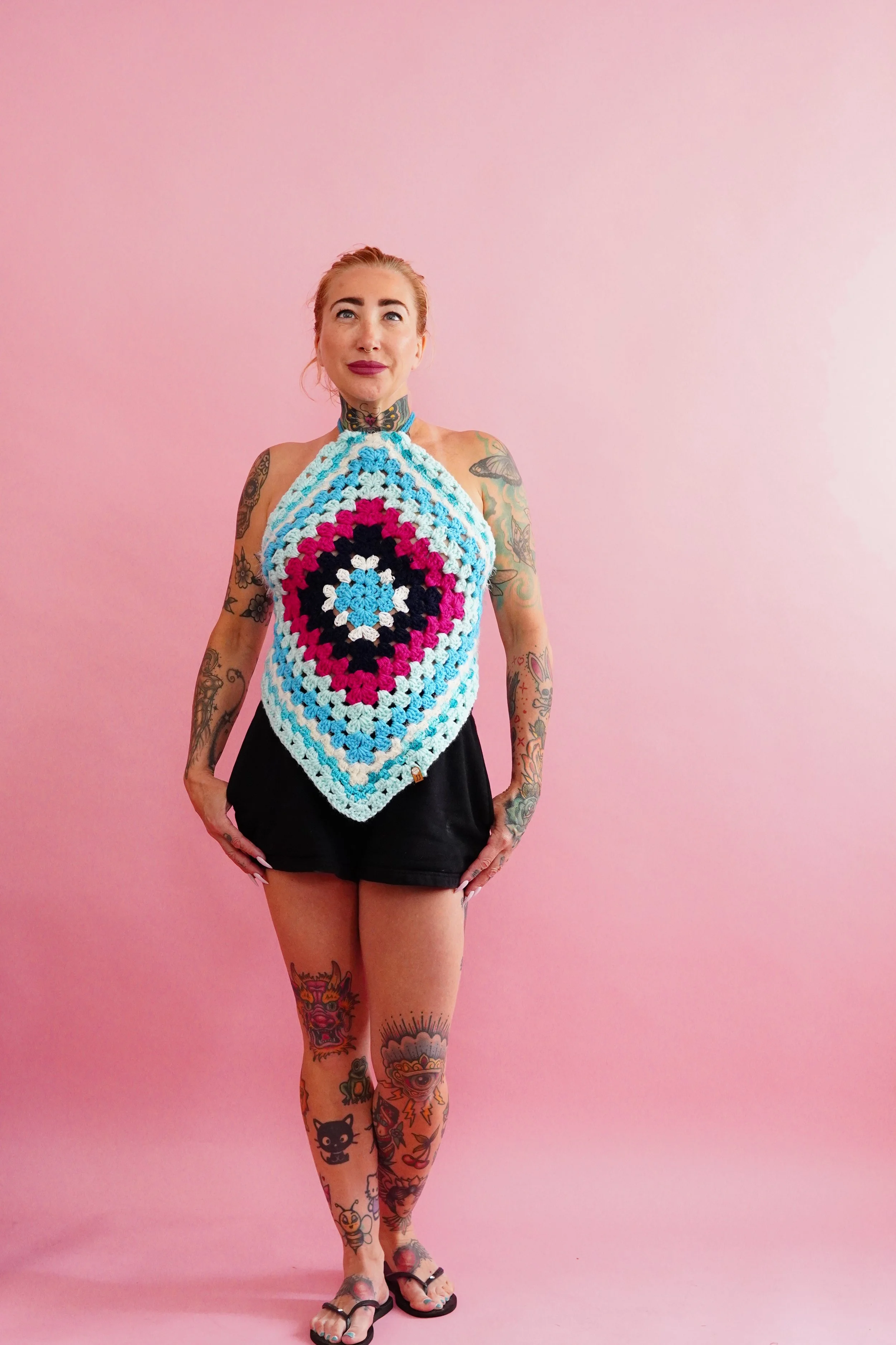 A woman with tattoos on her arms and legs standing against a pink background, wearing a colorful crocheted halter top and black shorts.
