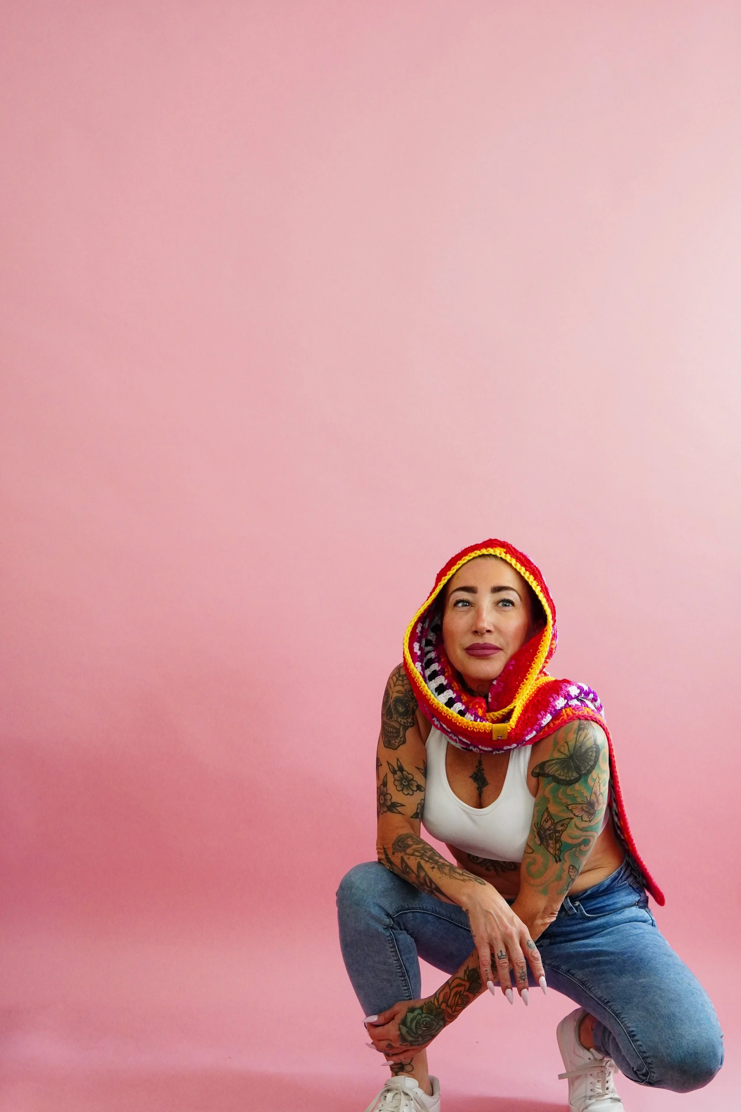 A woman with tattoos on her arms squats against a pink background, wearing a white tank top, light blue jeans, and a colorful crochet hooded scarf.