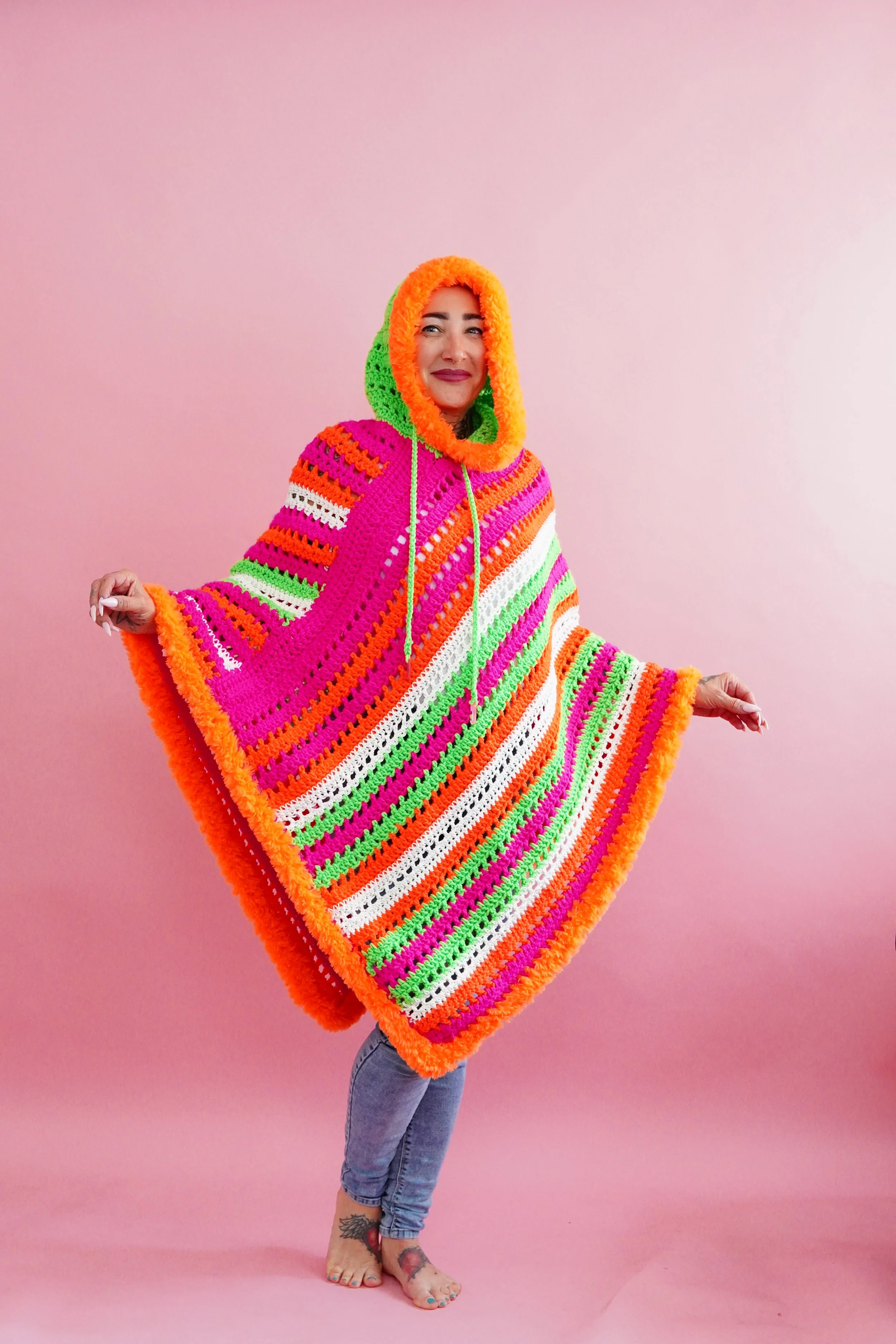 Woman wearing a colorful crochet glow-in-the-dark poncho with a hood, standing against a pink background, smiling.