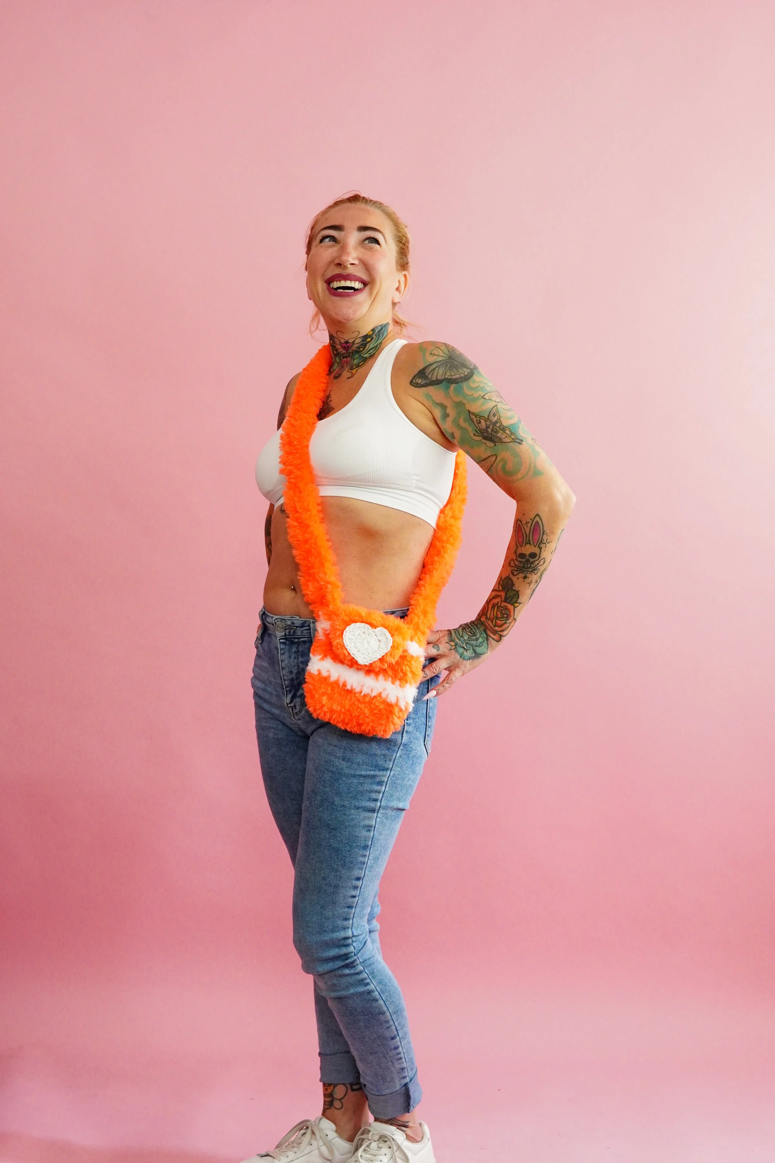A woman with tattoos wearing a white sports bra, jeans, white sneakers, and a handmade crochet orange fluffy crossbody bag, standing against a pink background and smiling.