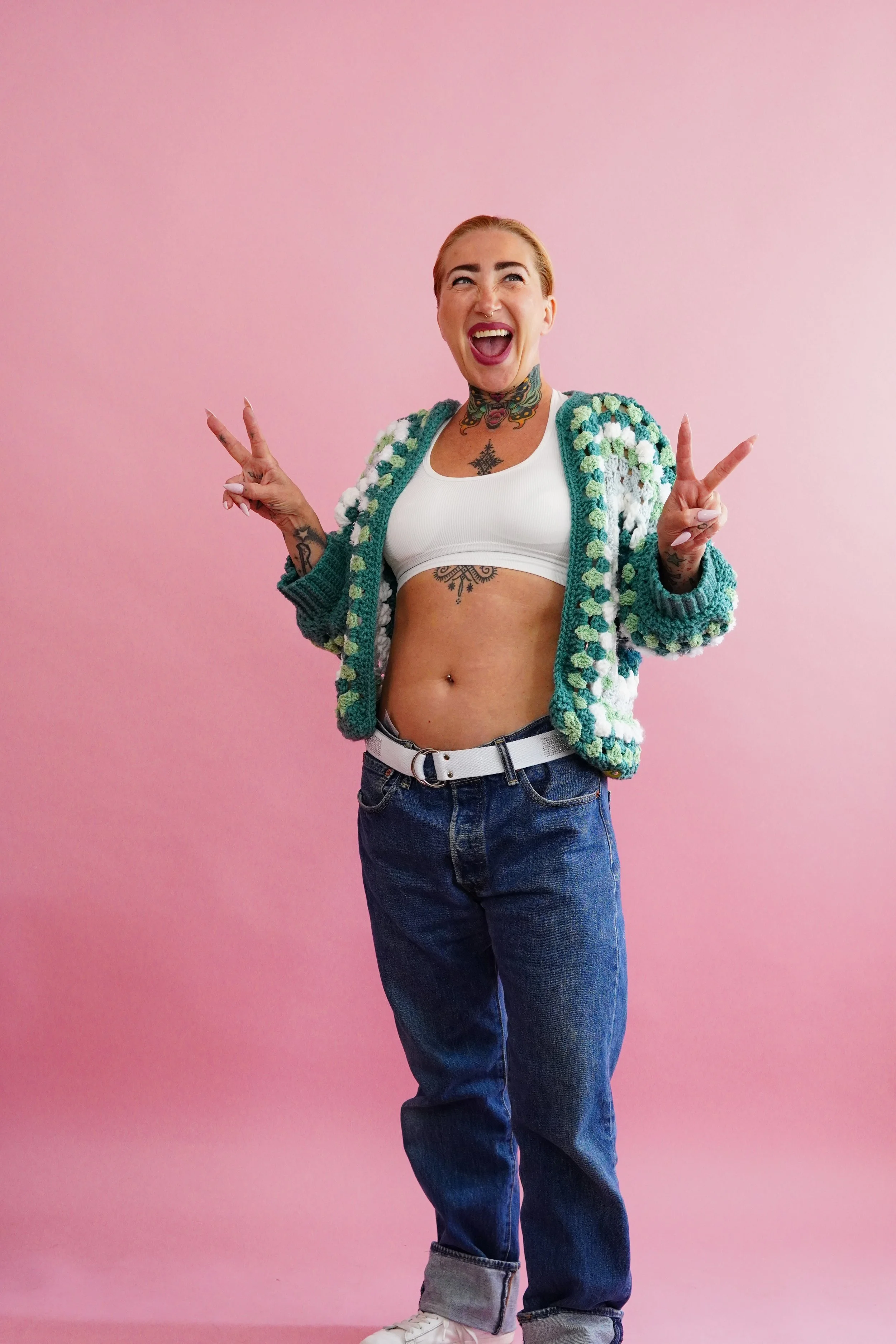 A woman with tattoos, wearing a white crop top and a colorful crocheted jacket, standing against a pink background. She is smiling, holding up peace signs with both hands.