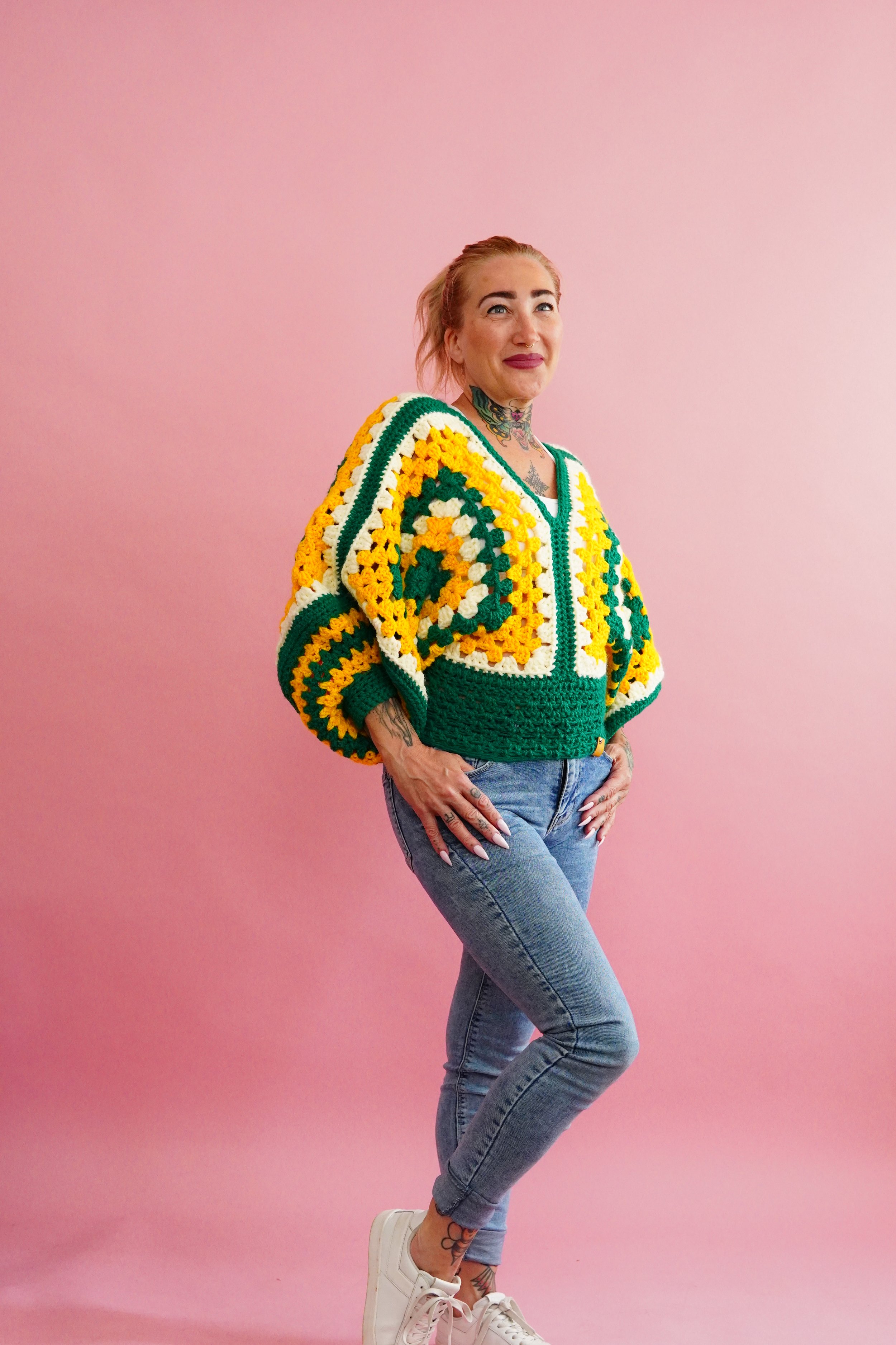 A woman with dyed pink hair standing against a pink background, wearing a colorful crochet sweater with green, yellow, white, and orange geometric patterns, blue jeans, and white sneakers.
