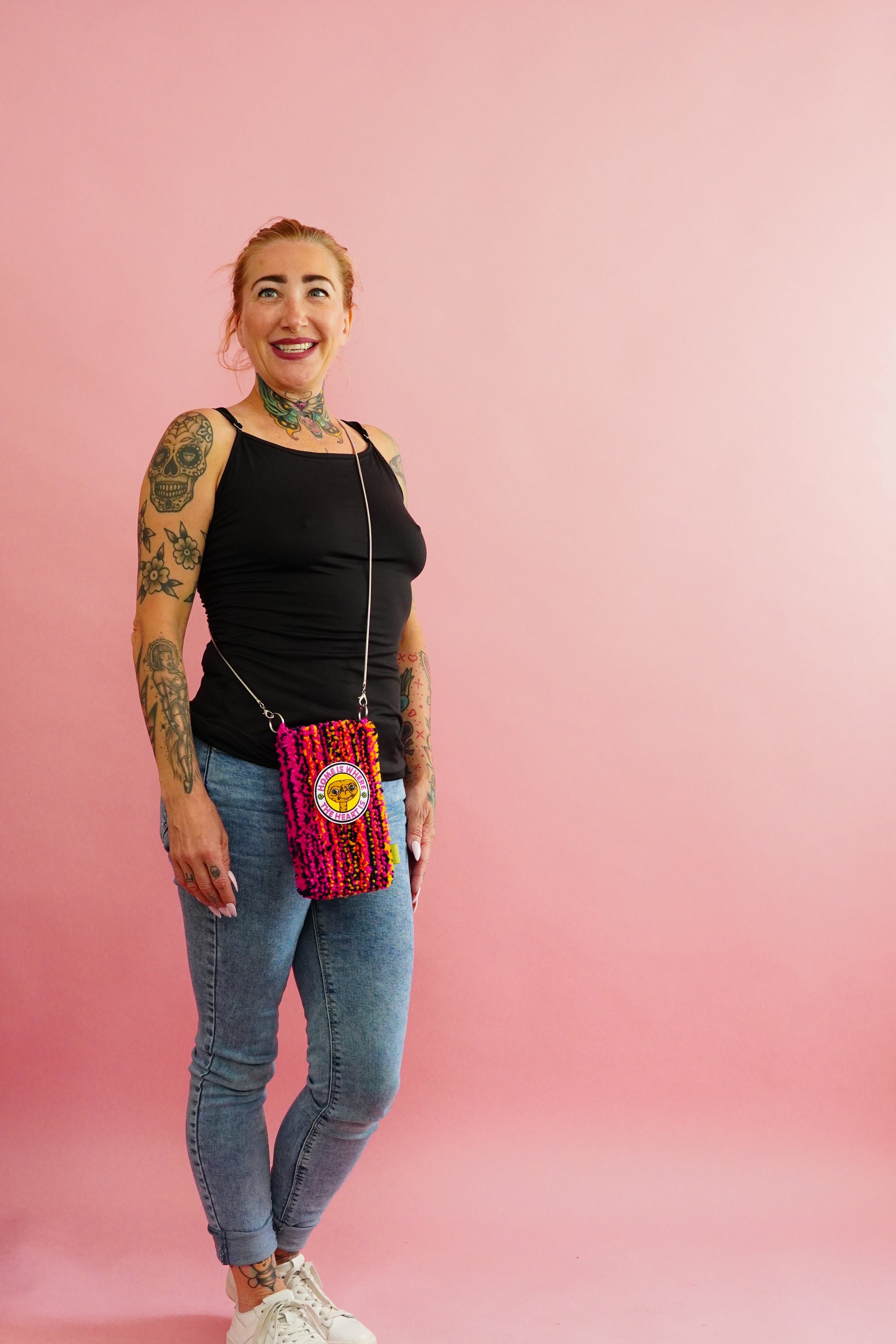 A smiling woman with tattoos on her arms and neck, wearing a black tank top, blue jeans, white sneakers, and carrying a colorful tufted rug bag with an E.T. patch, standing against a pink background.