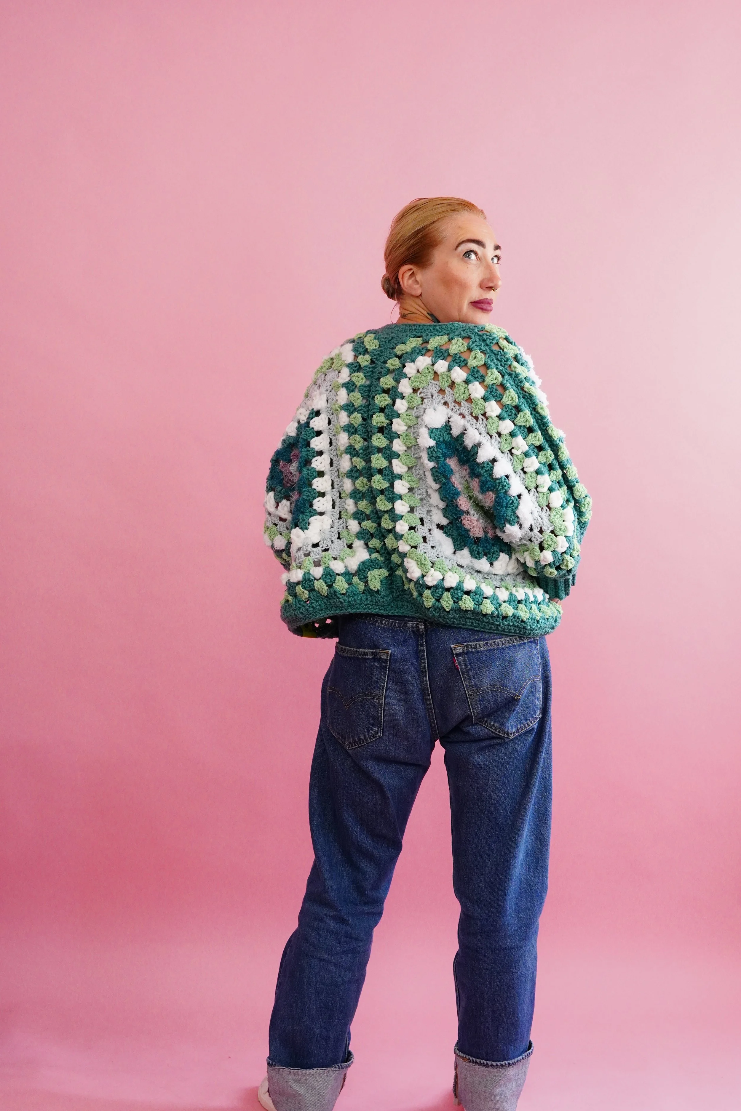 Woman with blonde hair in a braid, wearing a colorful crochet sweater, dark blue jeans with rolled cuffs, standing against a pink background, looking over her shoulder.