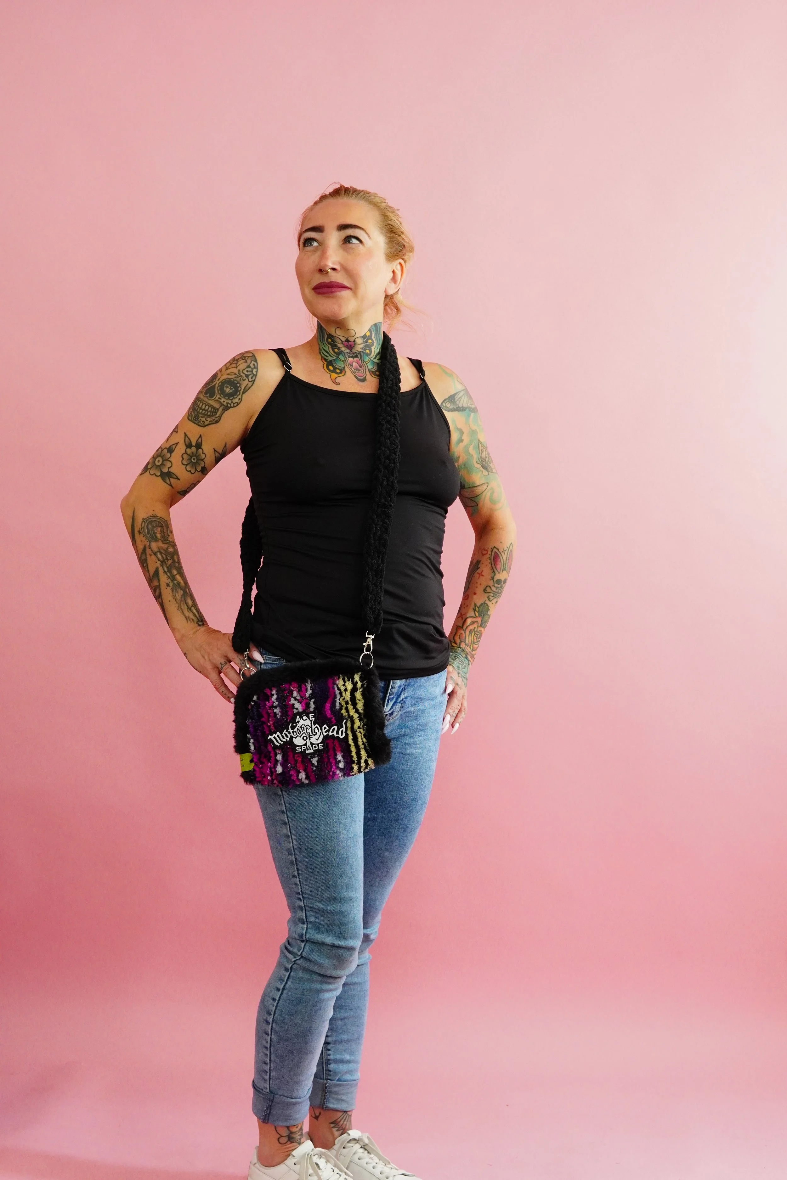A woman with multiple tattoos wearing a black tank top, light blue jeans, white sneakers, and a colorful fuzzy rug tufted purse, posing against a pink background.
