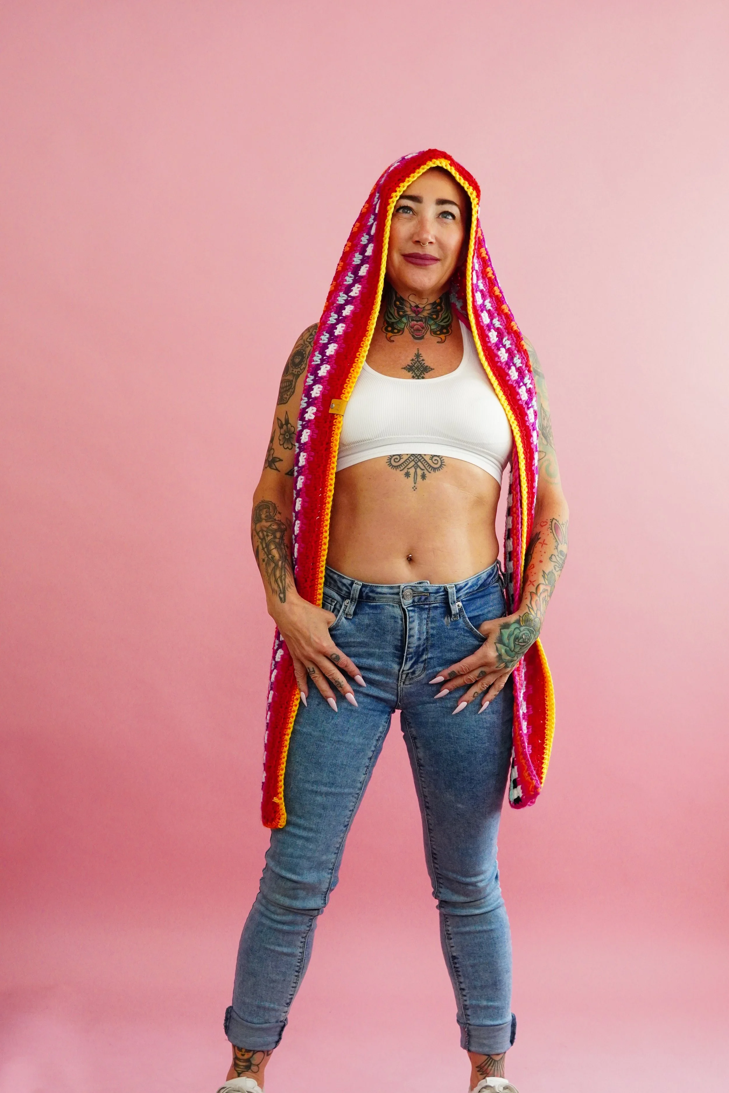 A woman with tattoos, wearing a white sports bra, blue jeans, and a colorful hooded crochet scarf on a pink background.