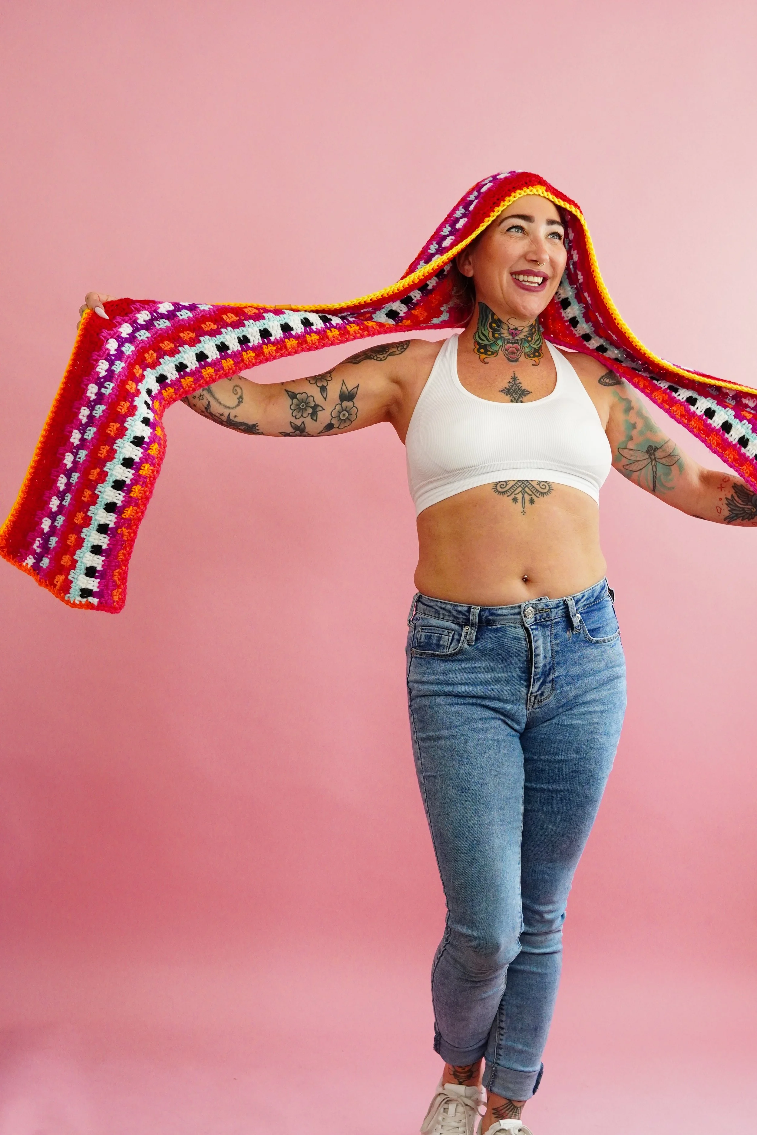 A woman with tattoos, wearing a white crop top and jeans, smiling while holding a colorful crocheted blanket over her head against a pink background.
