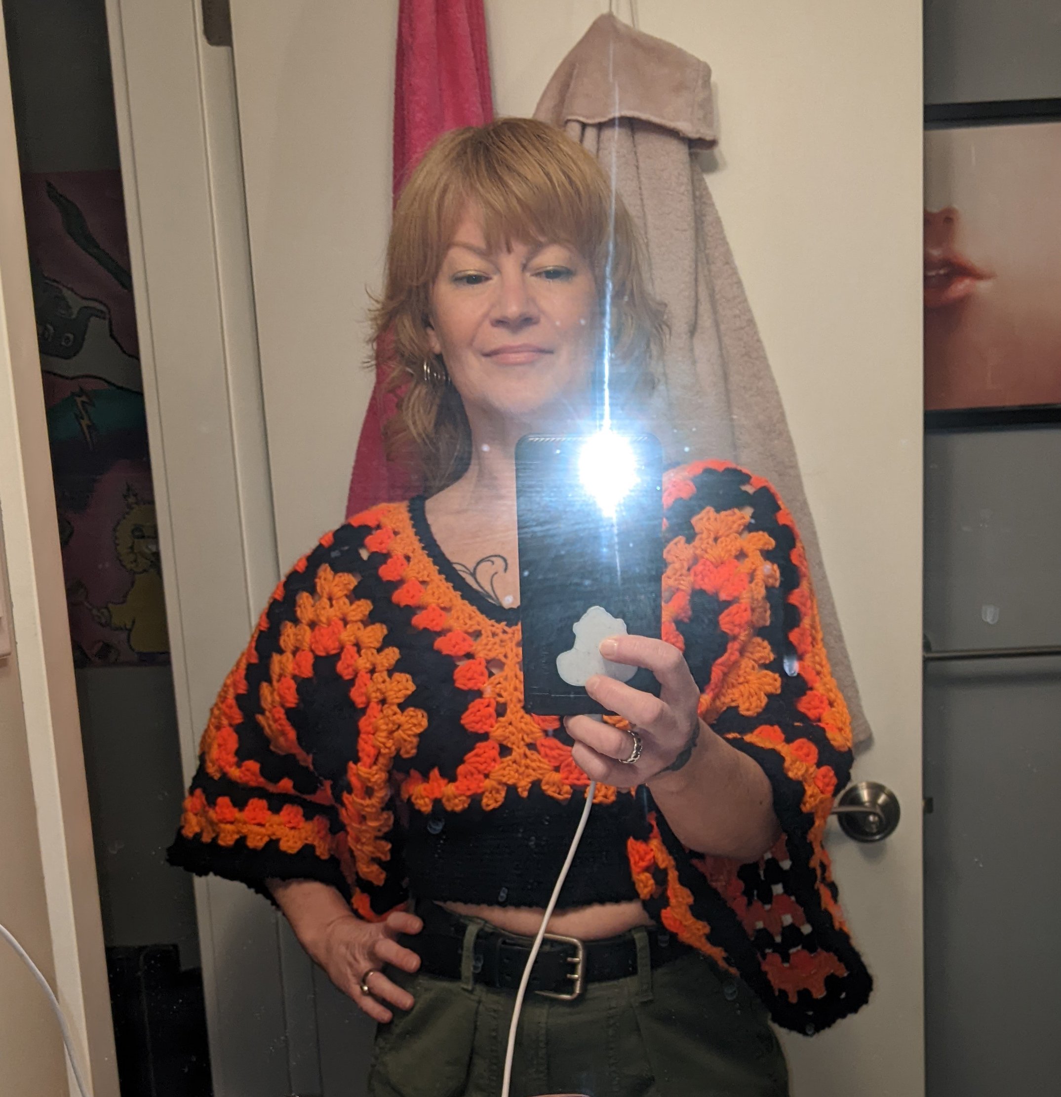 Woman taking a mirror selfie with her phone in a room. She has short reddish-blonde hair and is wearing a black and orange crochet sweater, with green pants and a black belt.