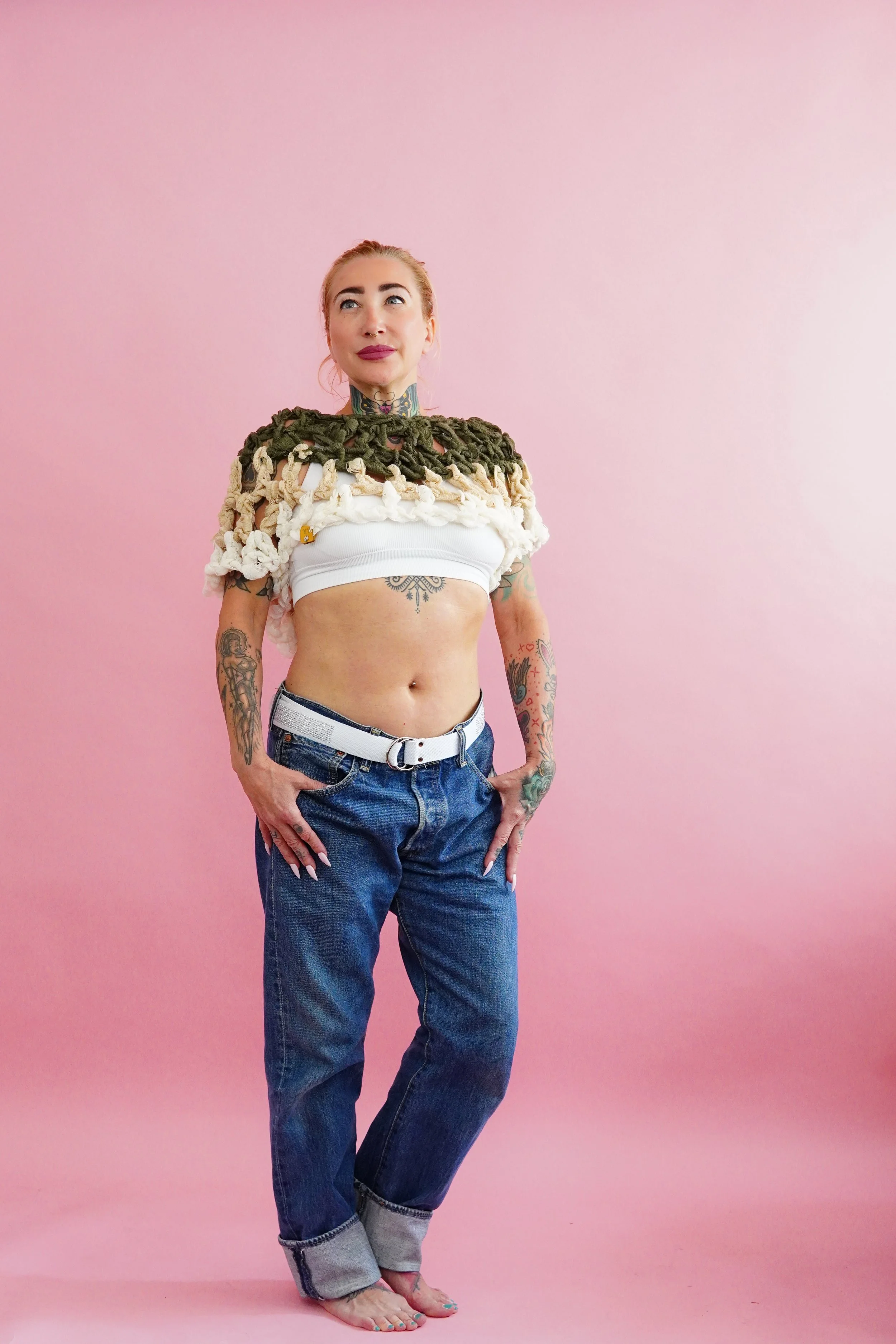 A woman with tattoos standing barefoot against a pink background, wearing jeans with rolled-up cuffs and a white crop top, with an intricate crochet handmade shawl around her shoulders.