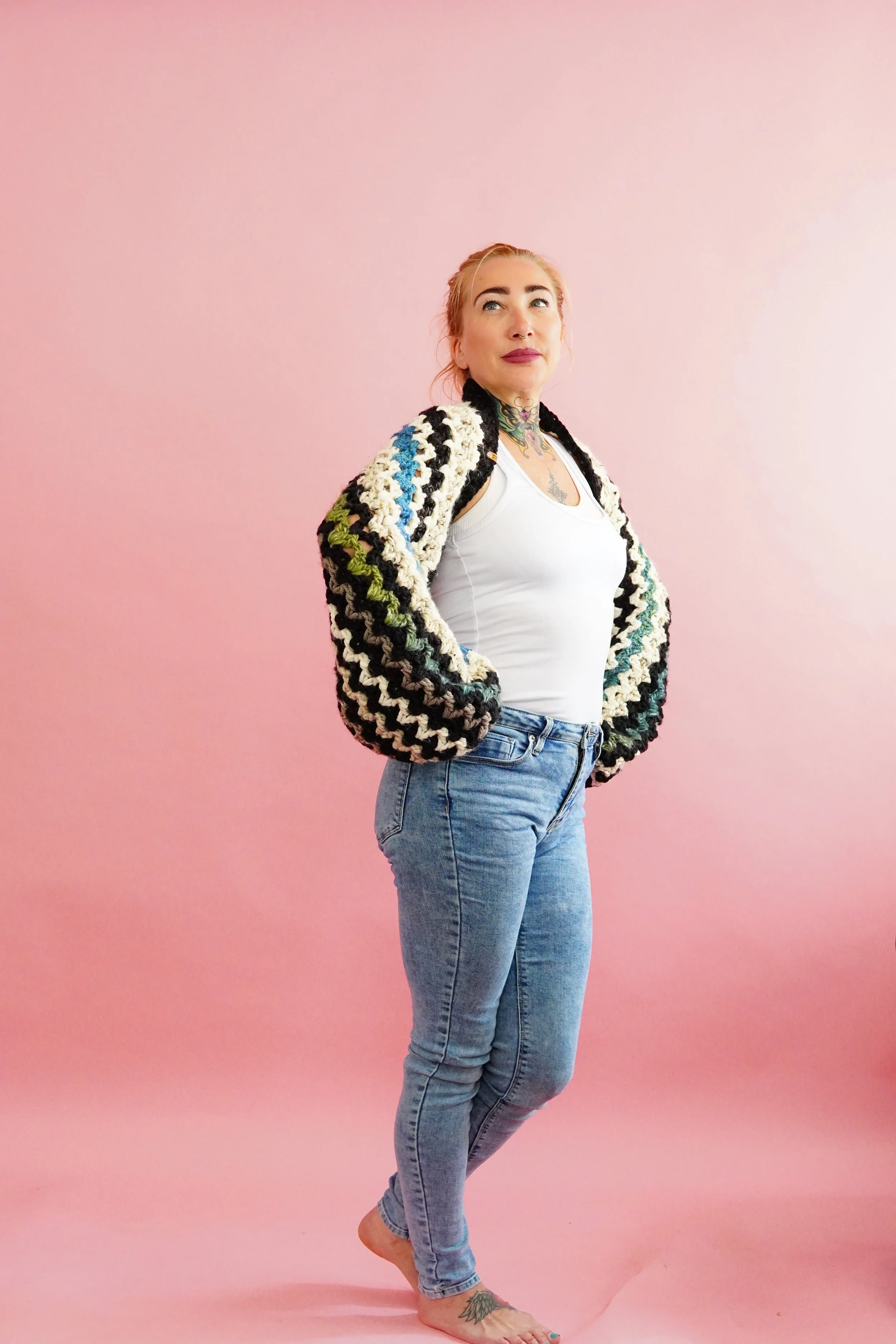 A woman with blonde hair and tattoos wearing a multicolored crocheted jacket, white tank top, and jeans standing barefoot against a pink background.